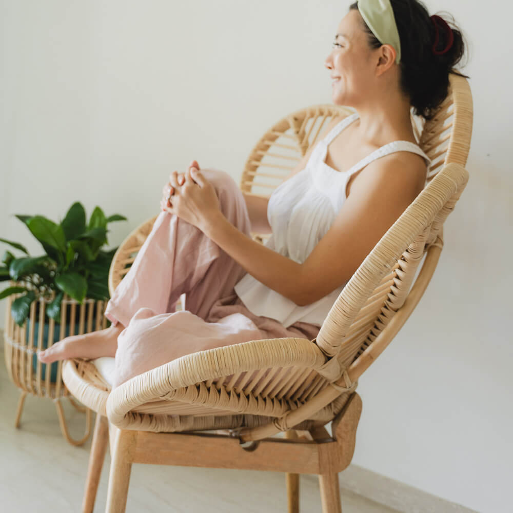 Side view of a woman sitting comfortably on a handcrafted wicker Petal Chair by MOMIJI in a cozy room with indoor plants.