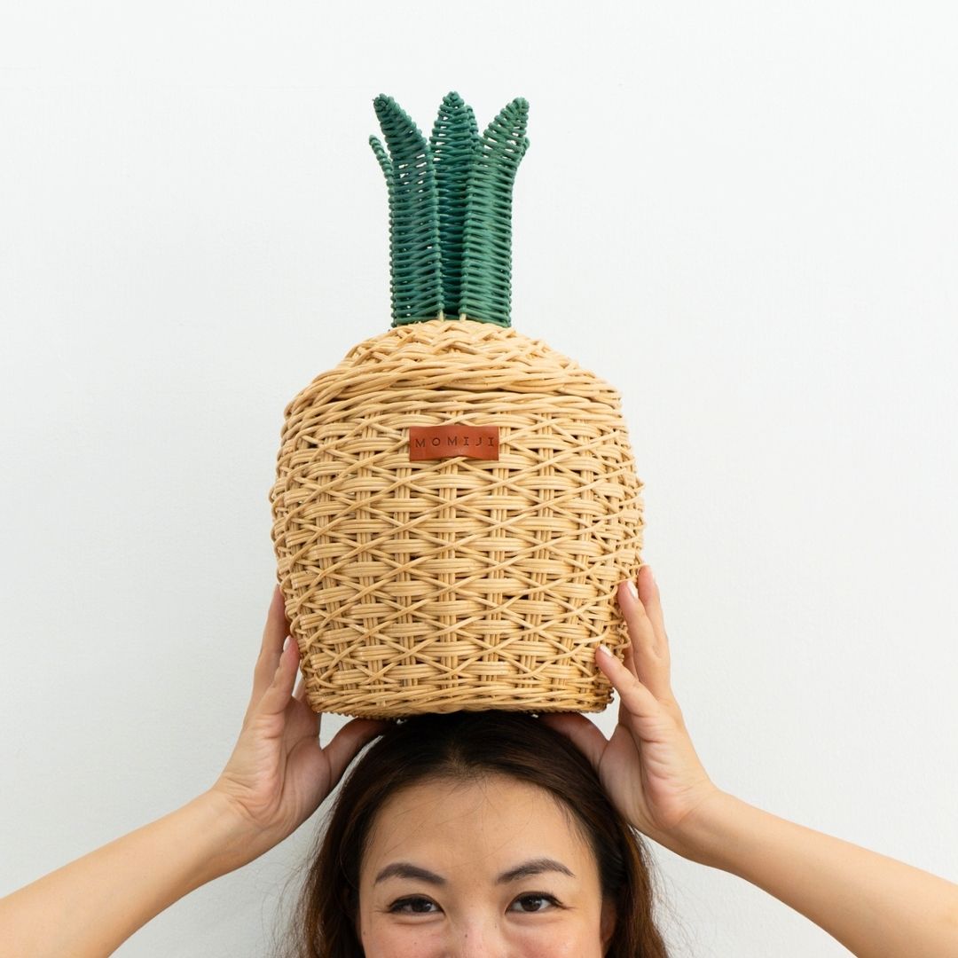 Woman holding a handcrafted natural pineapple rattan storage basket in small size by MOMIJI on her head against a white background.