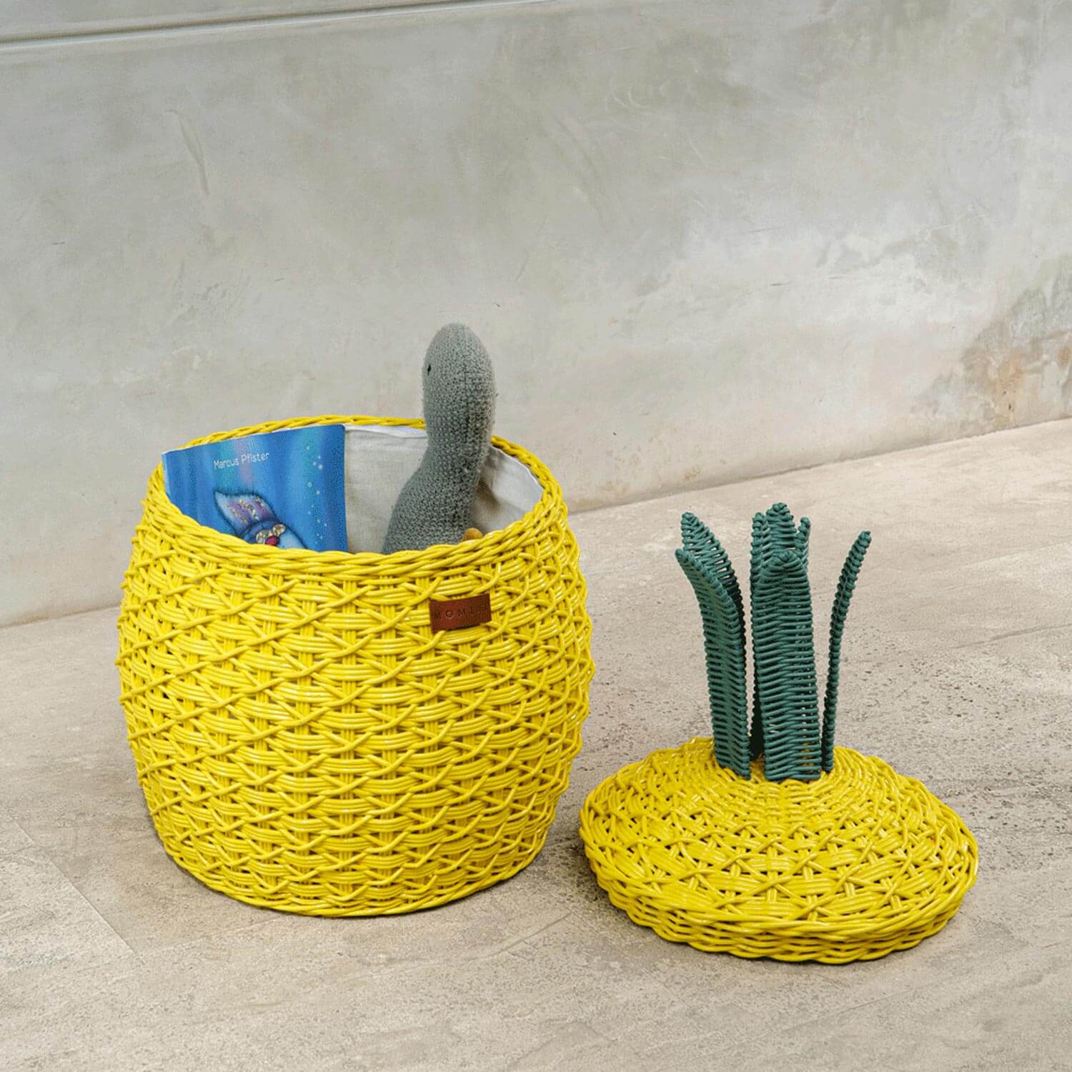 An opened yellow Pineapple Rattan Storage Basket with a book and a stuffed toy inside with lid on the floor.