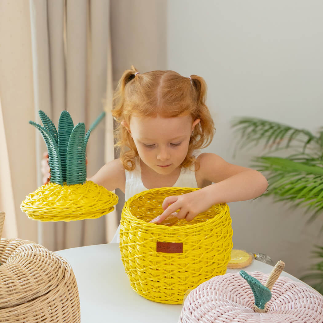 Child placing cookies into a handcrafted yellow pineapple rattan storage basket in small size by MOMIJI, styled with other fruit baskets on a table.