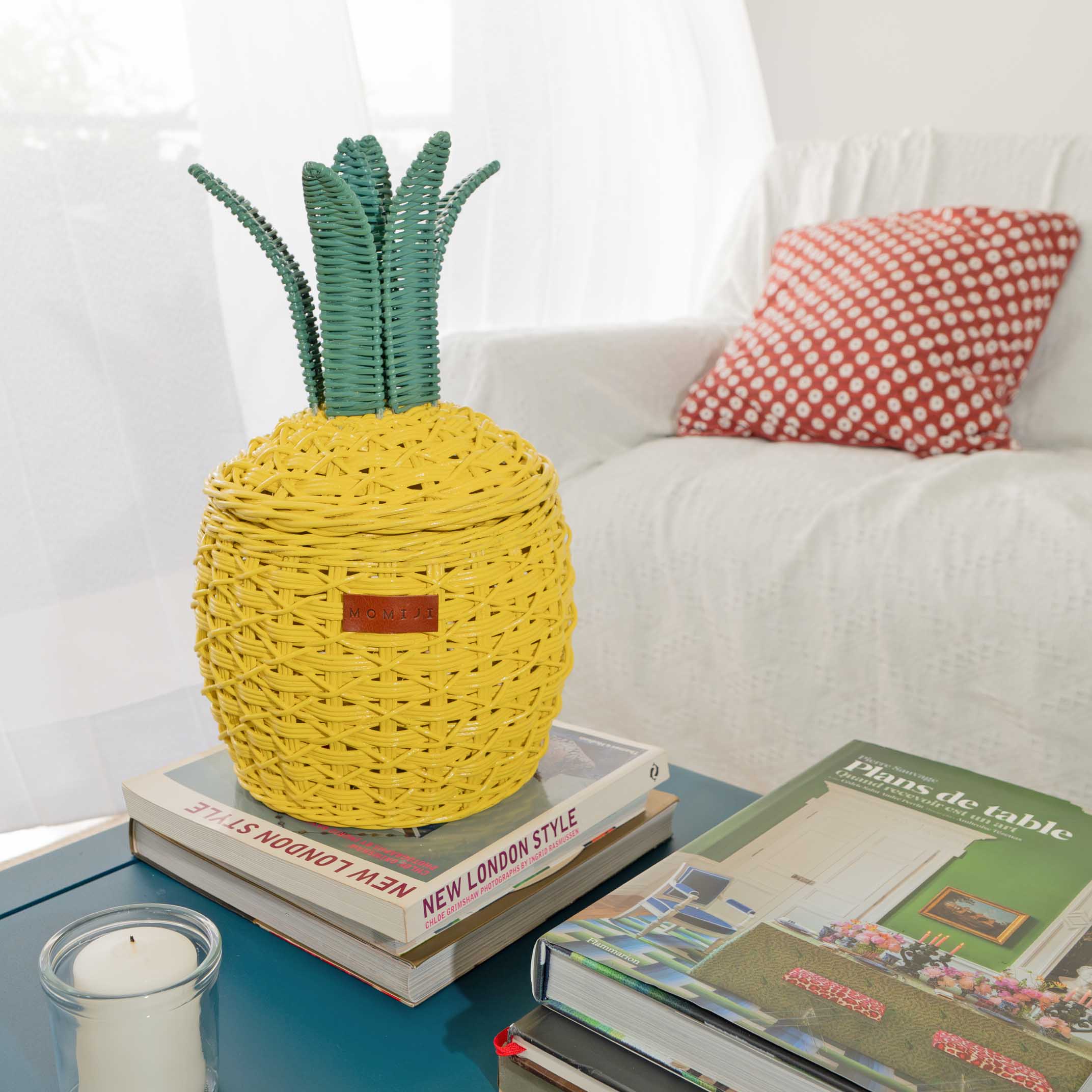 Handcrafted yellow Pineapple Rattan Storage Basket in small size by MOMIJI on books with a candle and magazine on a coffee table.