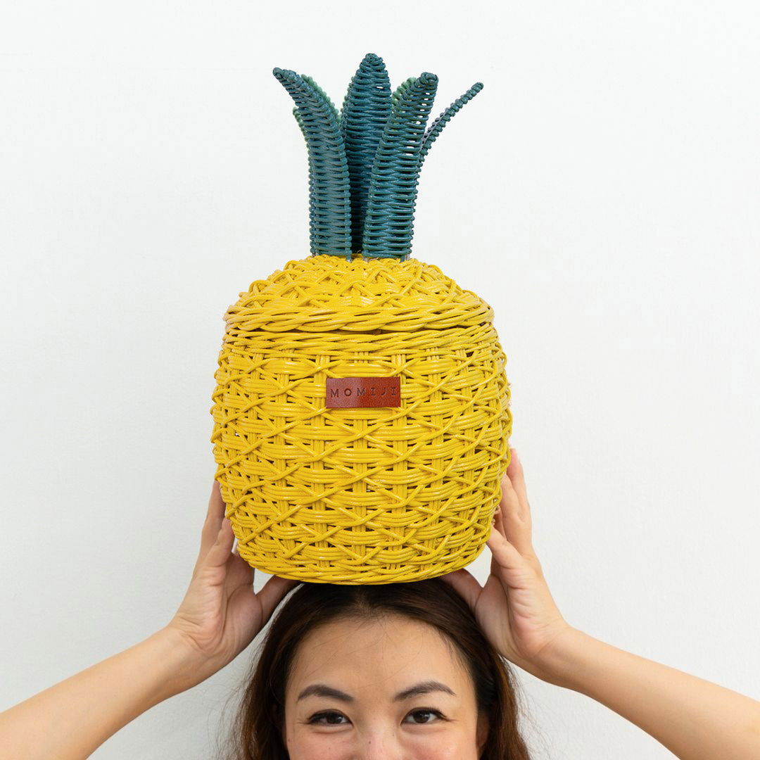 Woman holding a handcrafted yellow pineapple rattan storage basket in small size by MOMIJI on her head against a white background.