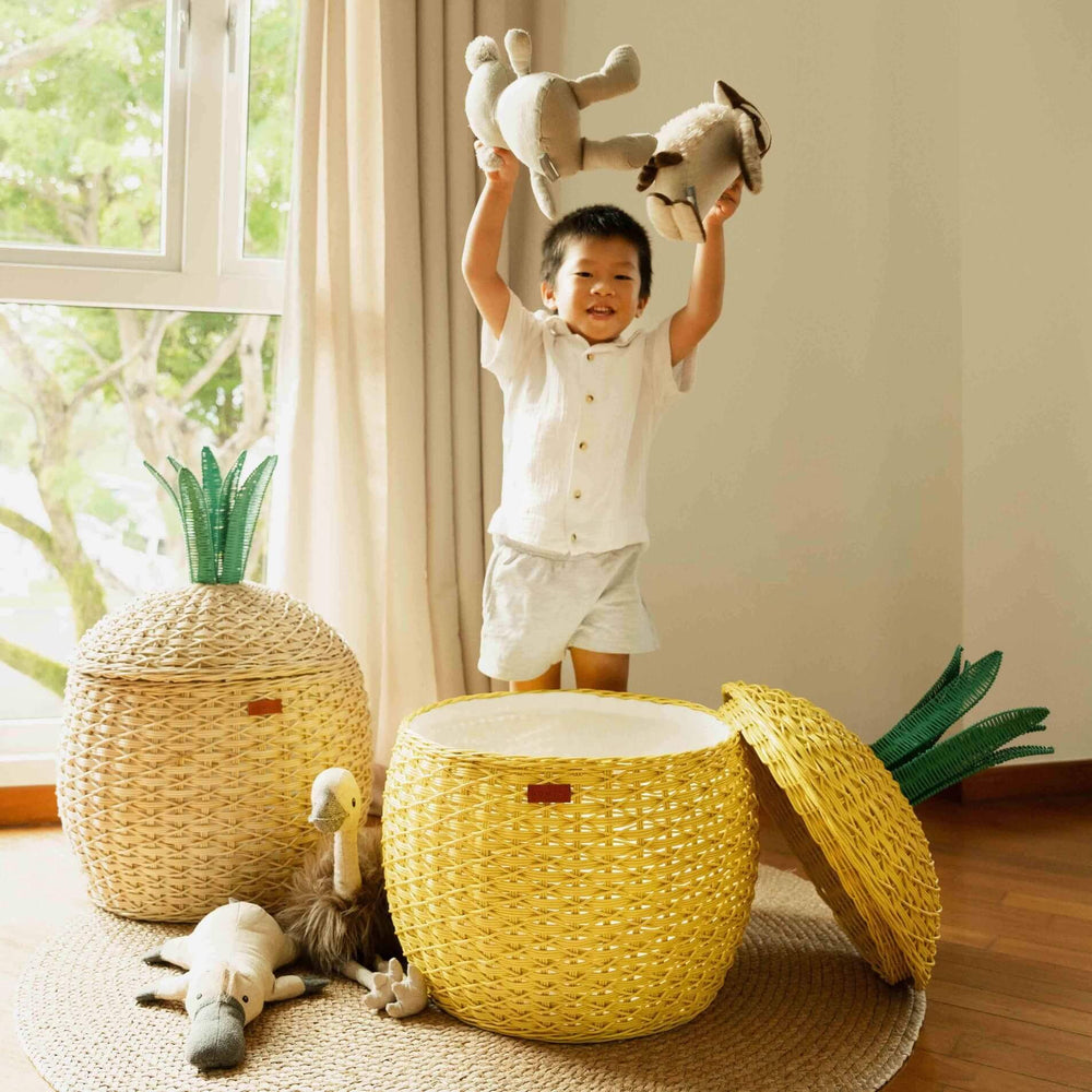 Child holding stuffed toys up high with yellow and natural colored Pineapple Rattan Storage Basket by MOMIJI, next to stuffed toys on the floor.