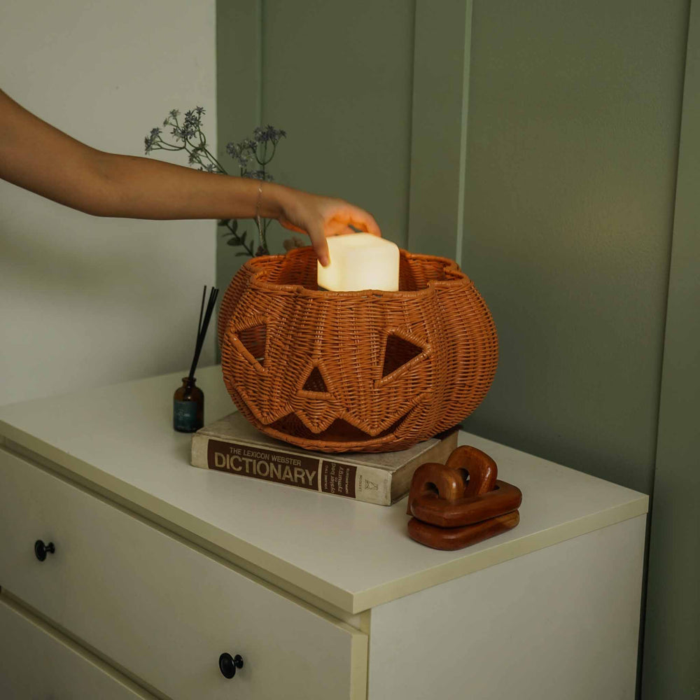 A hand placing a cube light into the handcrafted Pumpkin Face Rattan Basket by MOMIJI, showcasing its versatile design that transforms from a storage basket into a decorative lantern