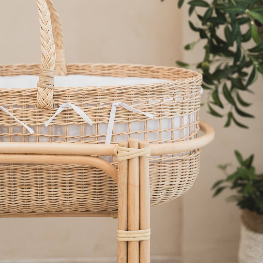 Wicker Moses basket with rocking stand by MOMIJI placed against a neutral wall with a plant in the background.