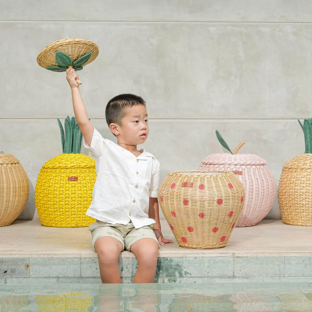Child holding a woven rattan basket lid by a pool with fruit designed rattan storage basket collection in the background.