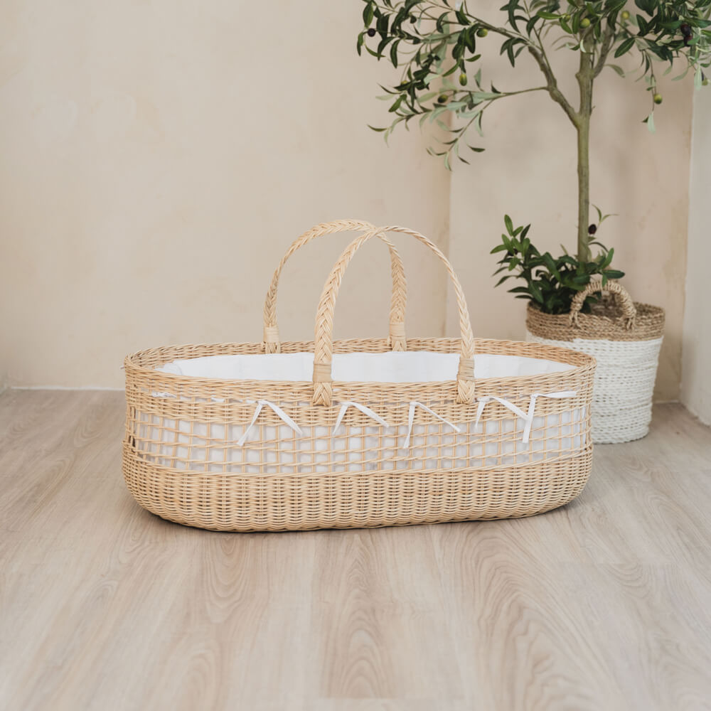 Vera Moses Rattan Basket by MOMIJI with a white mattress, placed on a wooden floor with a plant in the background, fully handwoven from natural rattan throughout the basket and handles.