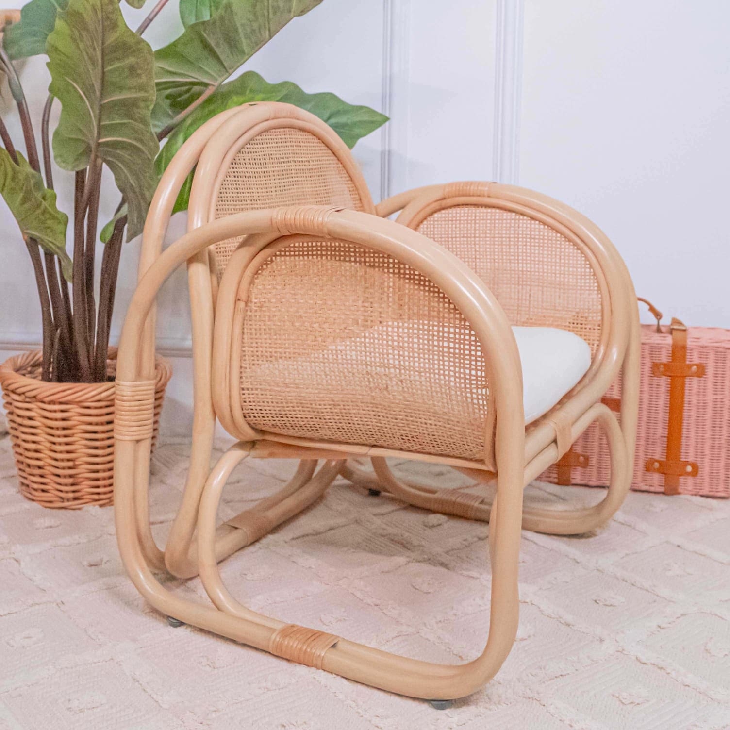 Side view of the child-sized rattan Willow Lounge Chair by MOMIJI with a white cushion on a wooden floor beside a plant and basket, showing the tightly woven armrest and reinforced side frame.