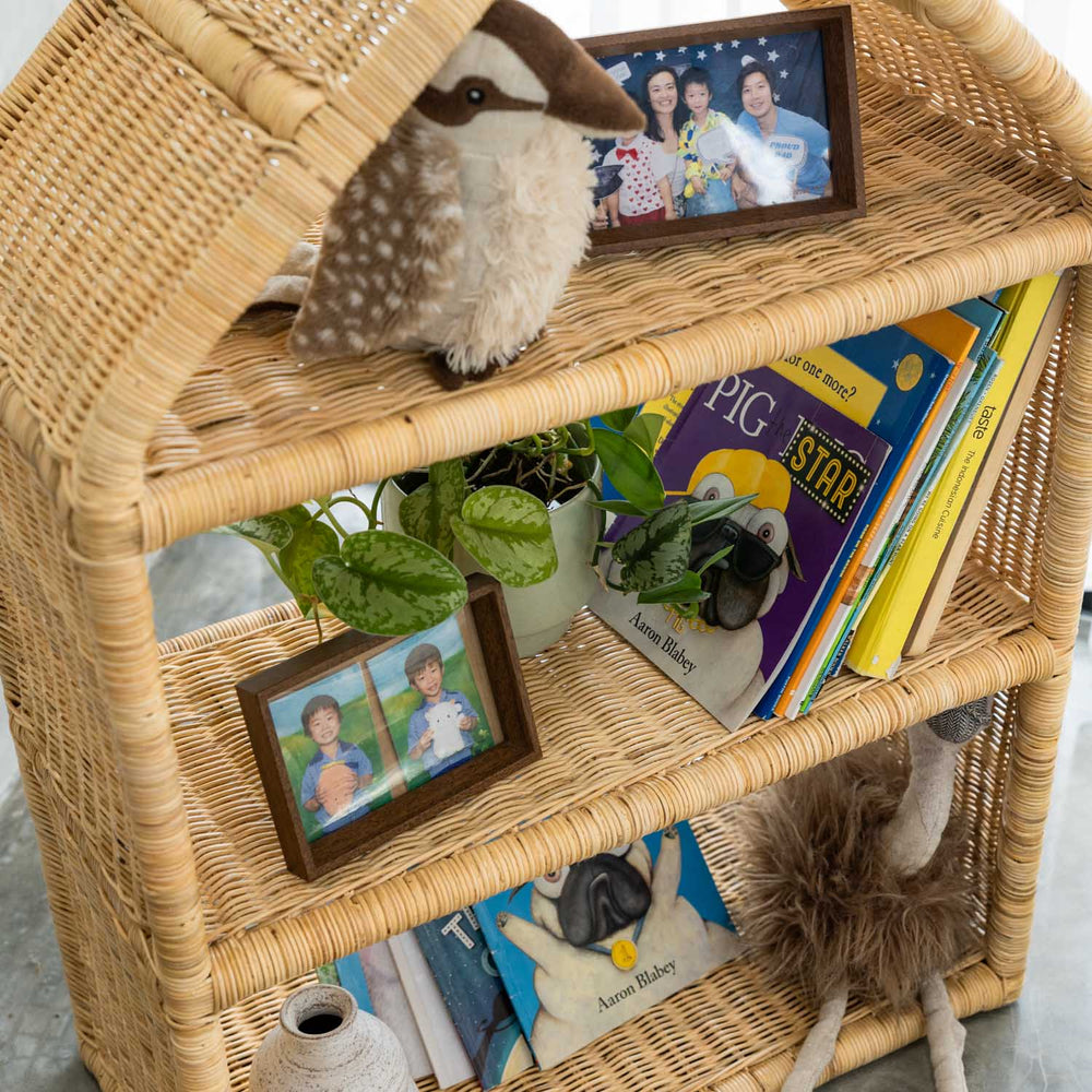 Top view of the handcrafted rattan wicker Alaya house shelf by MOMIJI, showing books, a stuffed animal, framed photos, and the fully woven rattan shelf surface.