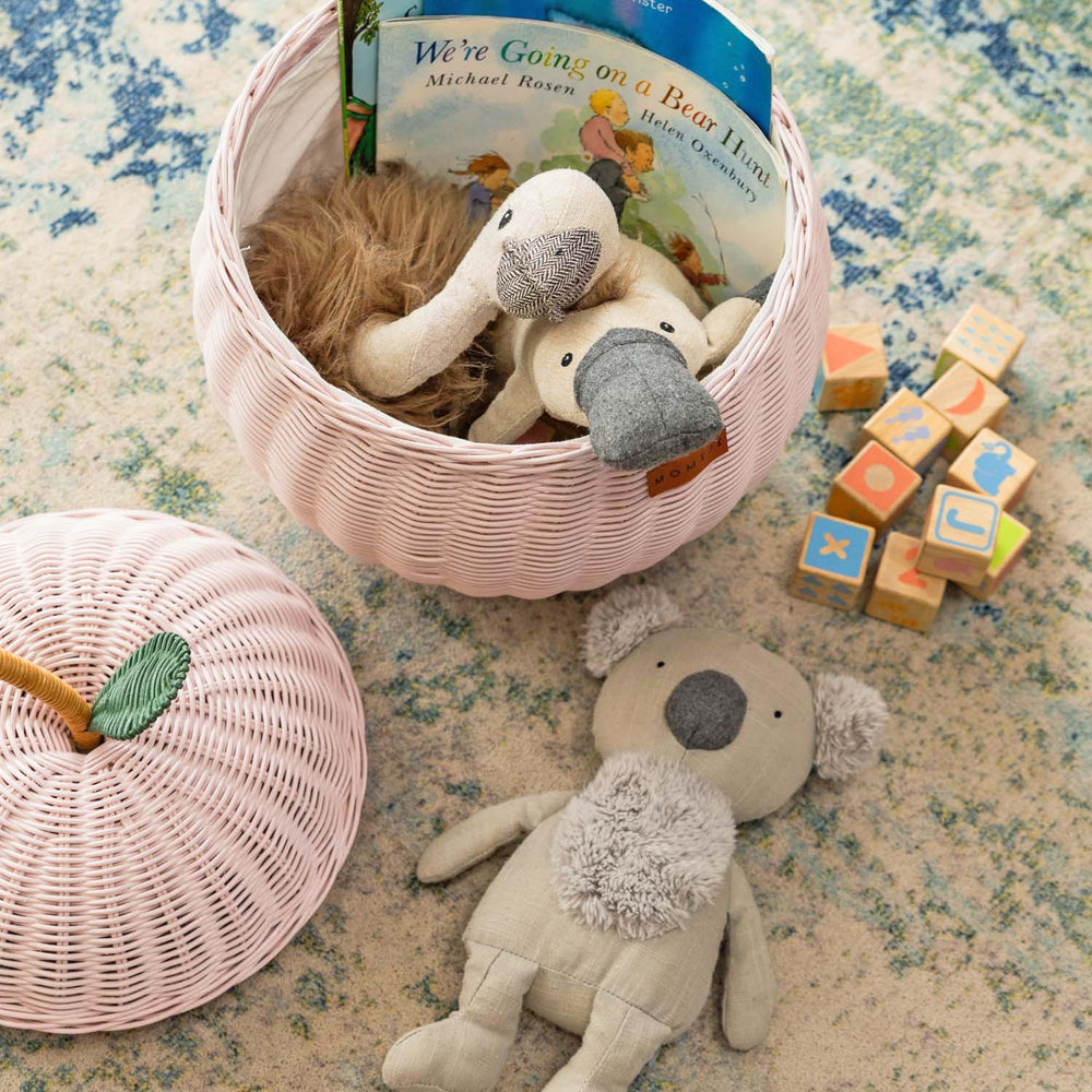 Handcrafted pink rattan apple storage basket in medium size by MOMIJI shown with the lid off, filled with stuffed toys, a book, and wooden blocks on a textured rug.