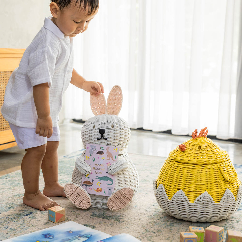 Child holding a handcrafted white rattan wicker bunny storage basket by MOMIJI in a room styled with a chick basket, toys, and children’s books.