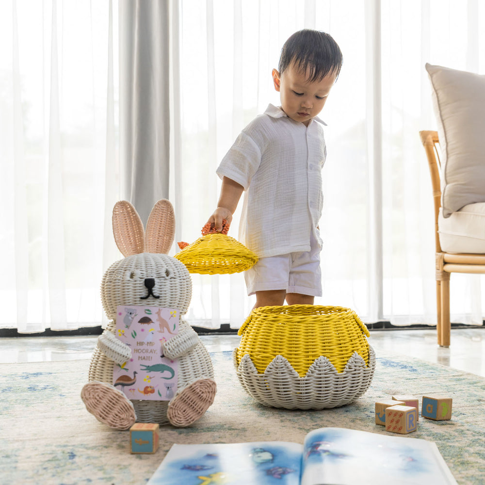 Child playing taking off the lid of handcrafted yellow rattan wicker chick basket by MOMIJI in a bright room, styled alongside a bunny rattan basket and children’s toys.