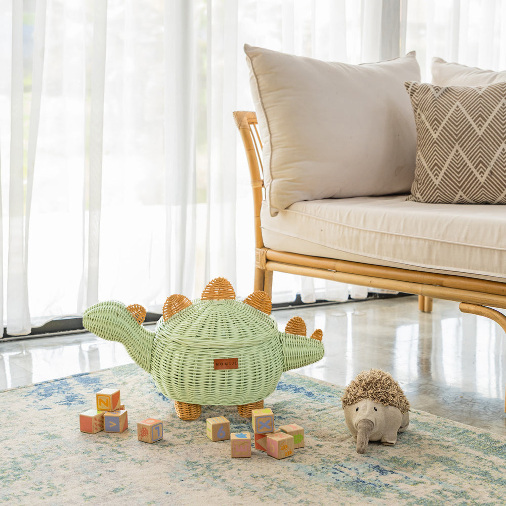 A medium size handcrafted dinosaur storage basket by MOMIJI in mint green rattan, styled with colorful blocks and a plush hedgehog toy on a rug in a kids room with a rattan bench.