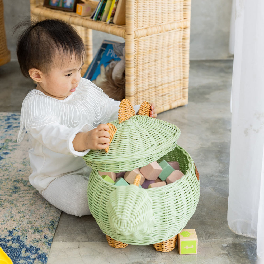 Child playing with a medium size handcrafted dinosaur storage basket by MOMIJI in mint green rattan, holding the lid open to reveal colorful blocks inside.