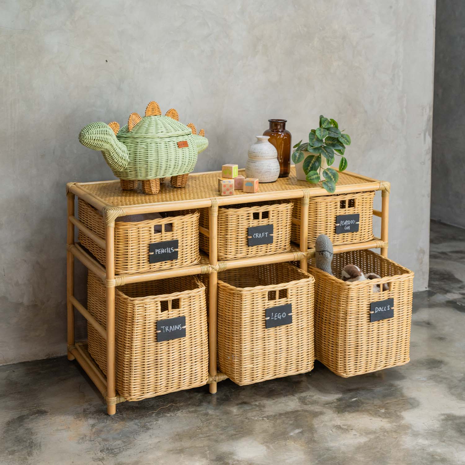 Graha Montessori rattan storage shelf by MOMIJI with blackboard-labeled baskets against a concrete wall, styled with a mint green rattan dinosaur basket and decor, with an open basket of stuffed toys.