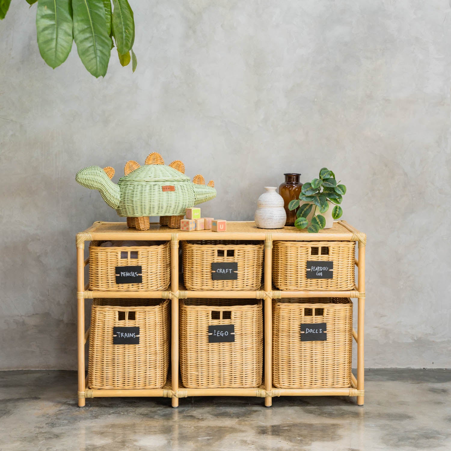 Graha Montessori rattan storage shelf by MOMIJI with blackboard-labeled baskets against a gray wall, styled with a mint green rattan dinosaur basket, a vase, and color block toys on top.
