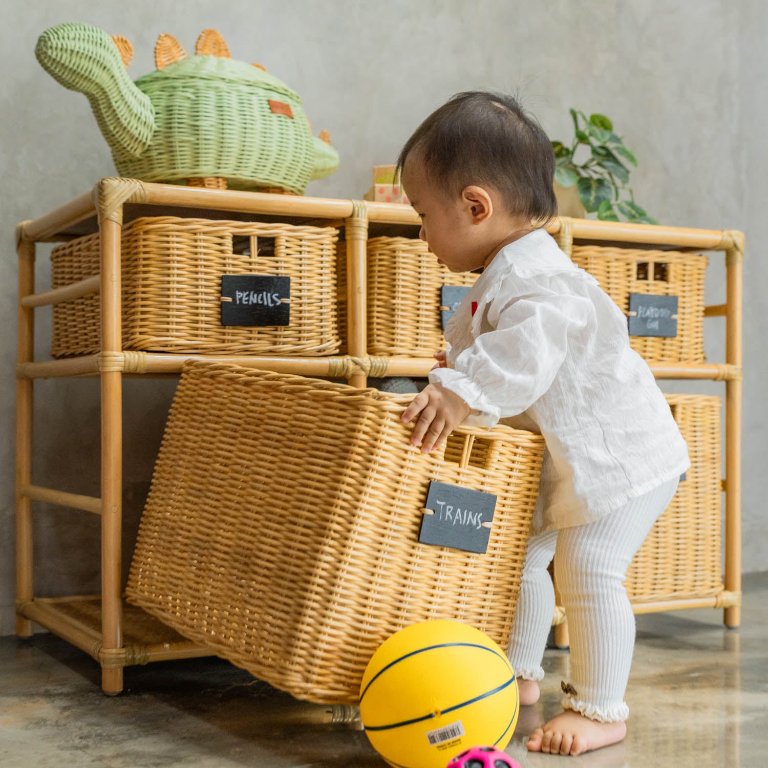 Child placing a blackboard-labeled “Trains” basket back onto the Graha Montessori rattan storage shelf by MOMIJI.