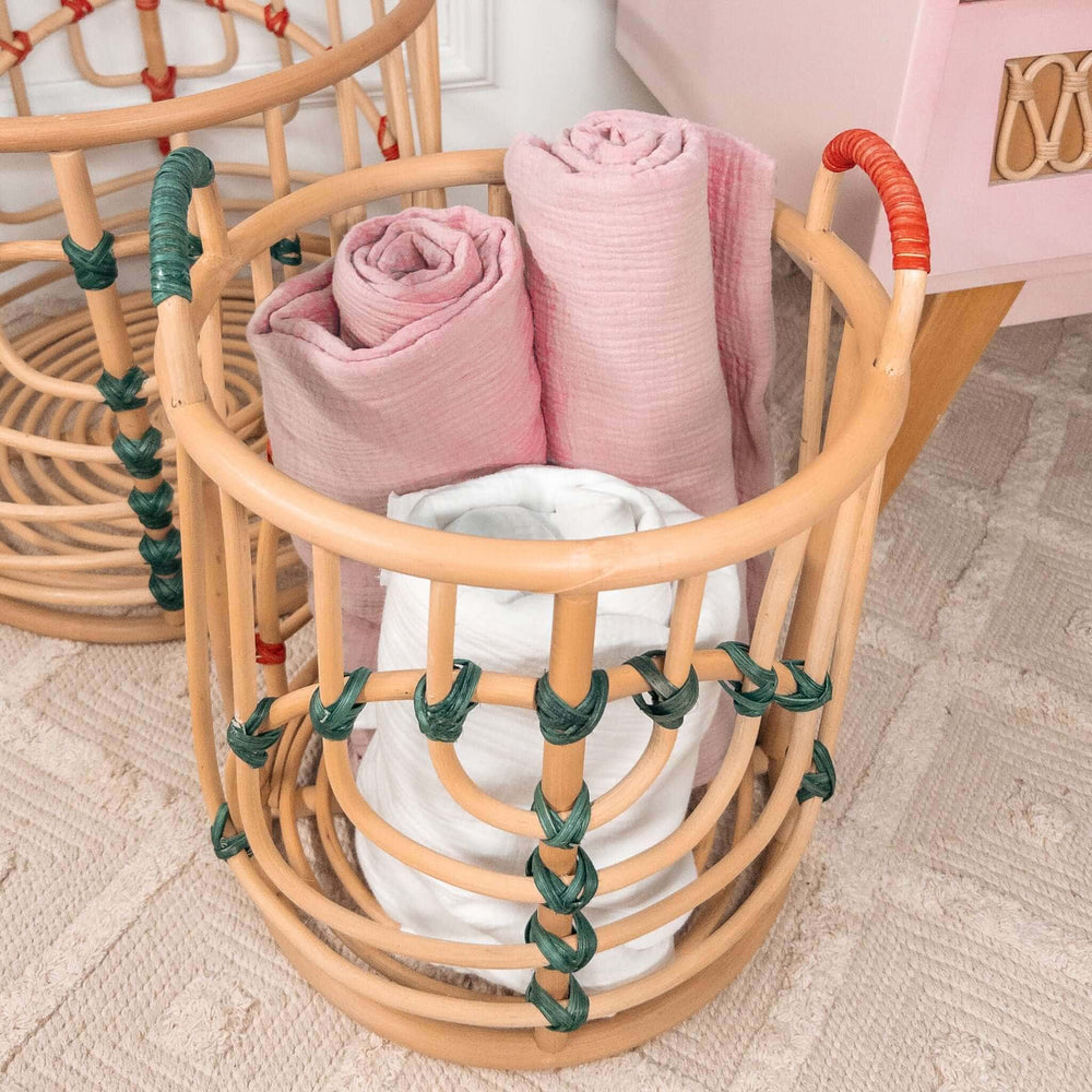 Close-up of the smaller handcrafted Iris baskets set by MOMIJI in natural rattan with red and green bindings filled with rolled towels, with the smaller basket beside a cabinet.