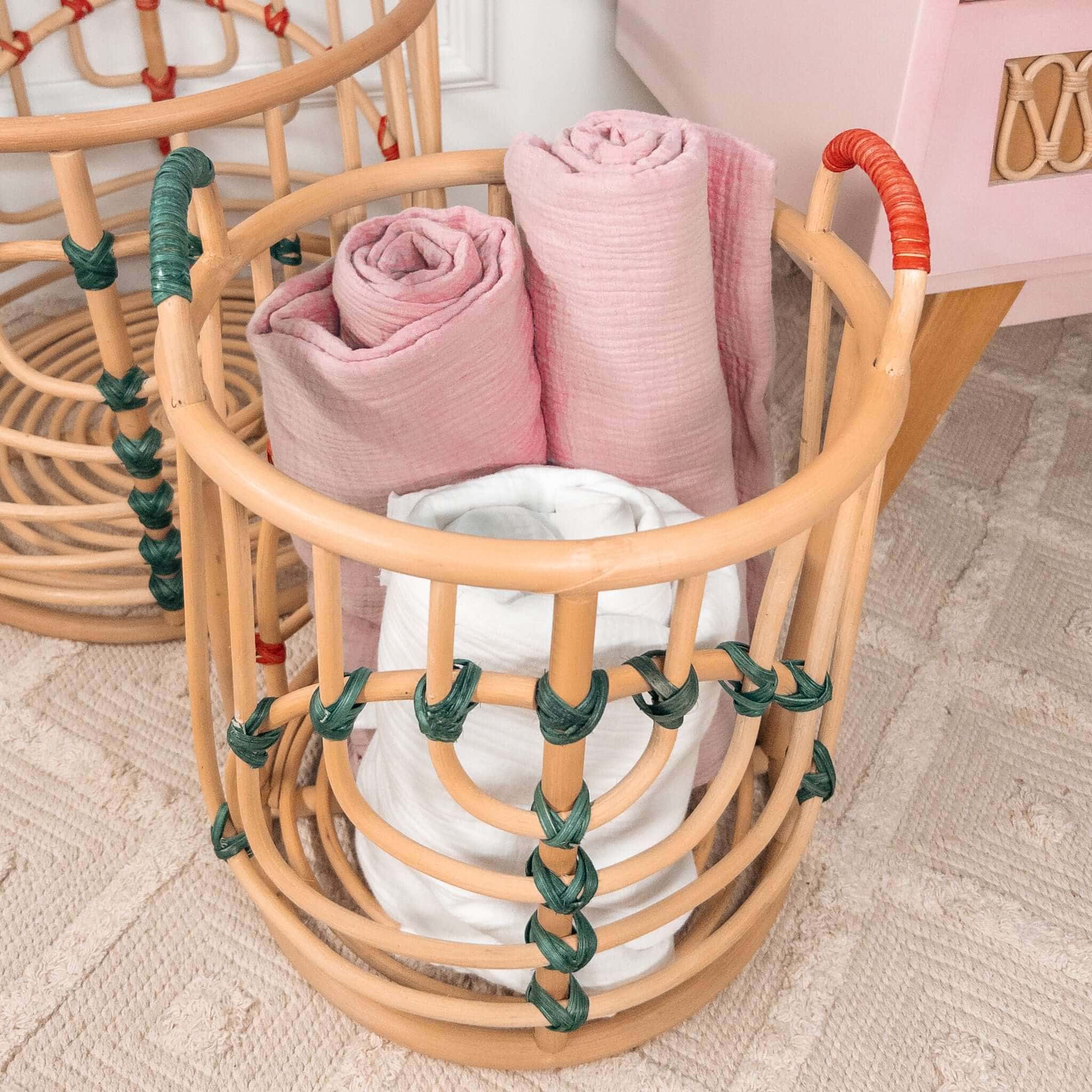 Close-up of the smaller handcrafted Iris baskets set by MOMIJI in natural rattan with red and green bindings filled with rolled towels, with the smaller basket beside a cabinet.