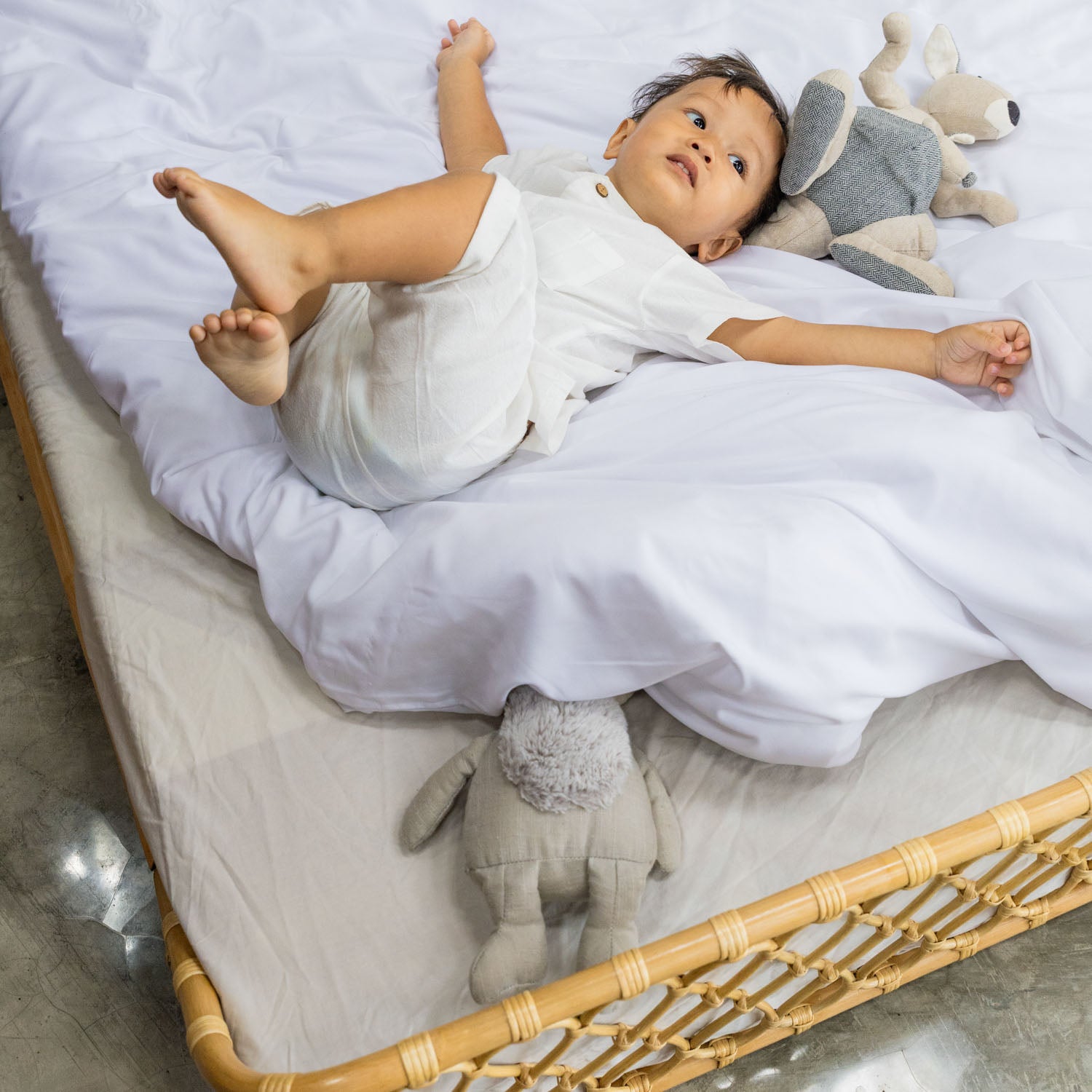 Baby lying on a white blanket with stuffed animals inside the handcrafted Layla Montessori rattan floor bed by MOMIJI in natural beige.