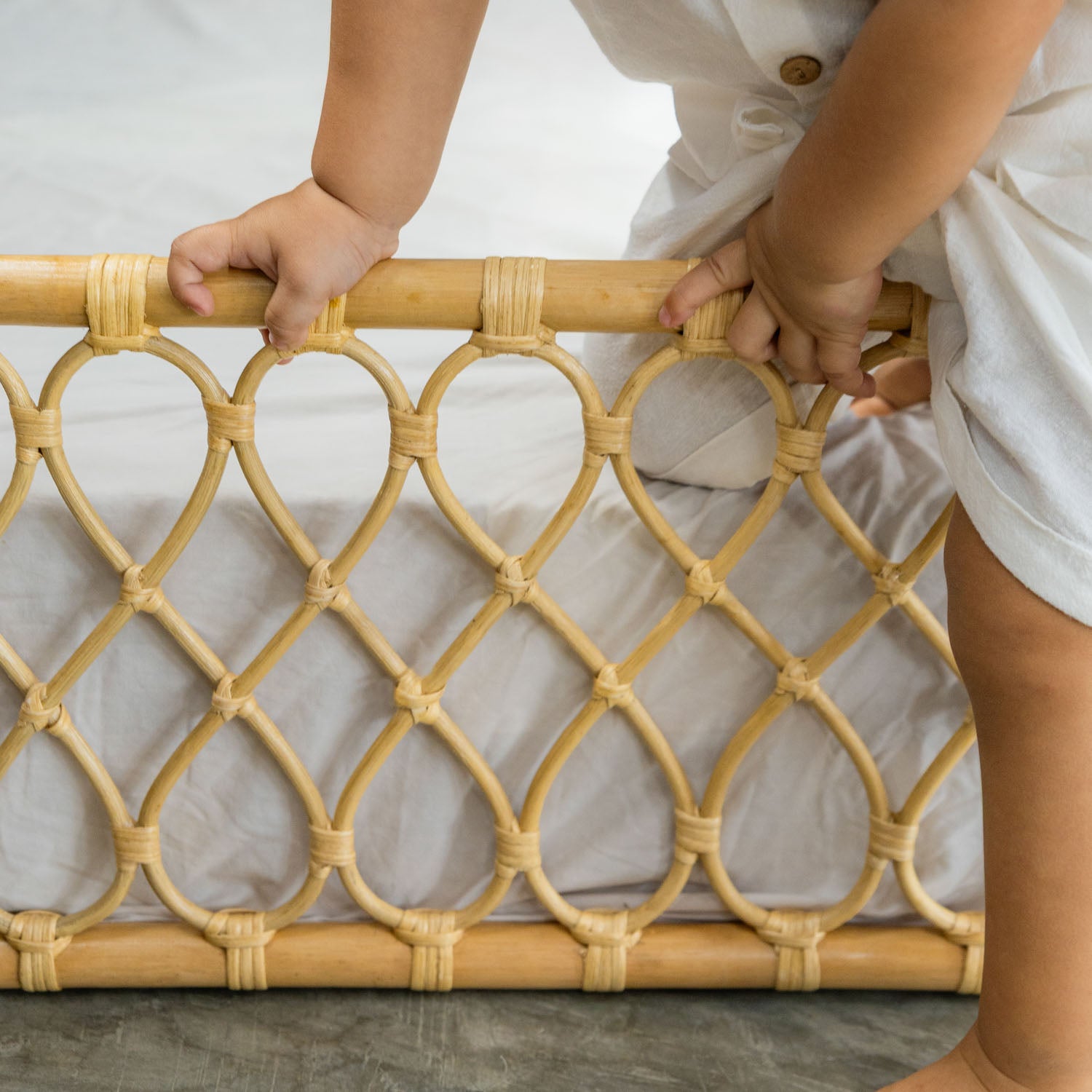 Child holding the carved frame of the handcrafted Layla Montessori rattan floor bed by MOMIJI in natural beige, showcasing the woven rattan craftsmanship.