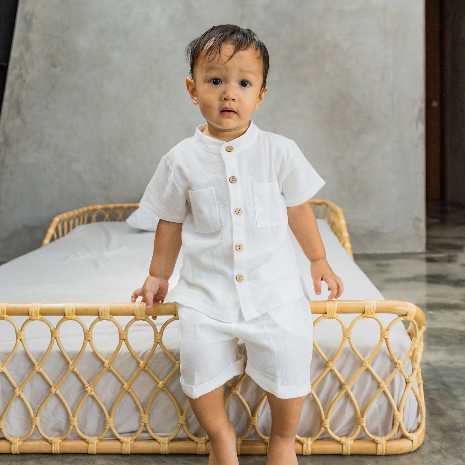 Child in a white outfit standing and leaning against the sturdy frame of the handcrafted Layla Montessori rattan floor bed by MOMIJI in natural beige.