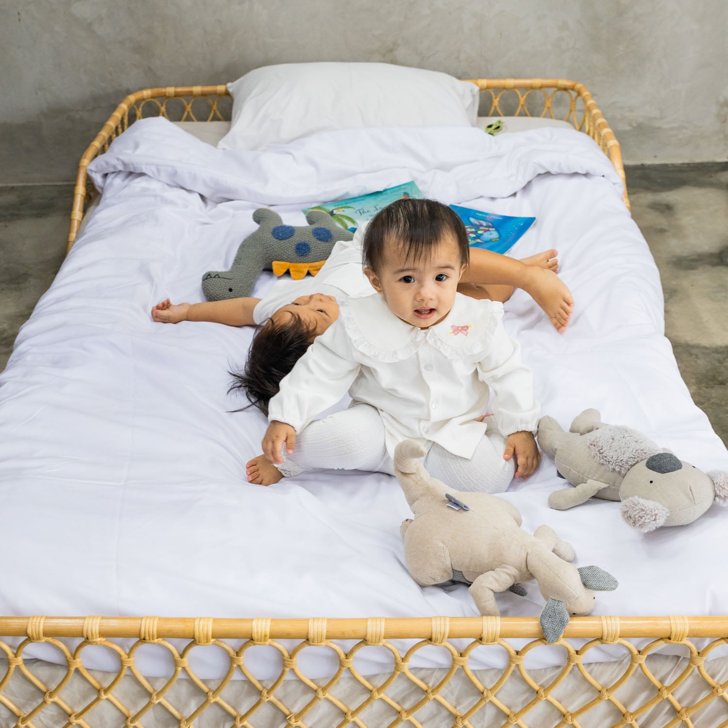 Two children on the handcrafted Layla Montessori rattan floor bed by MOMIJI in natural beige, styled with white bedding, stuffed animals, and children’s books.