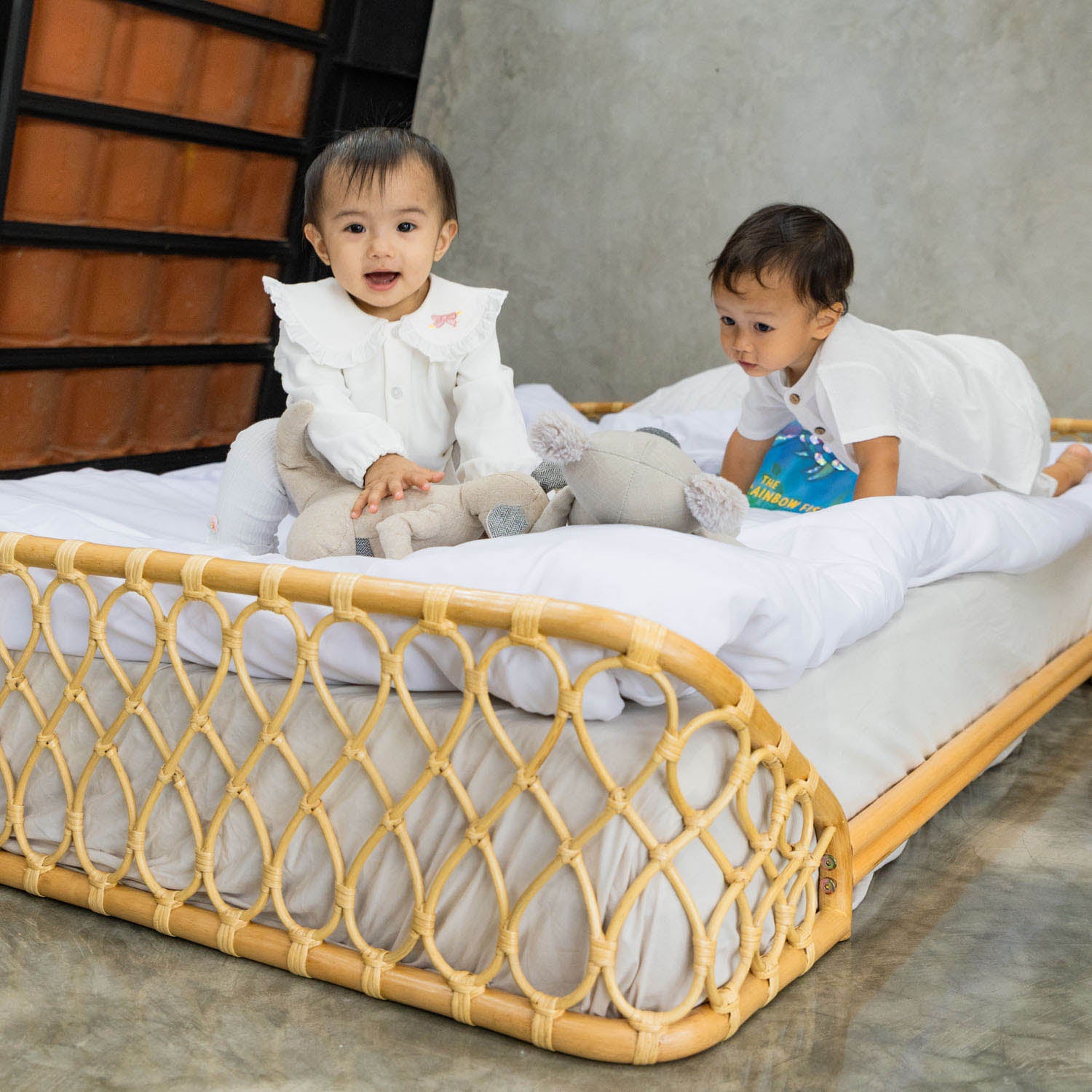 Two children sitting on the handcrafted Layla Montessori rattan floor bed by MOMIJI in natural beige, styled with white bedding and stuffed toys.