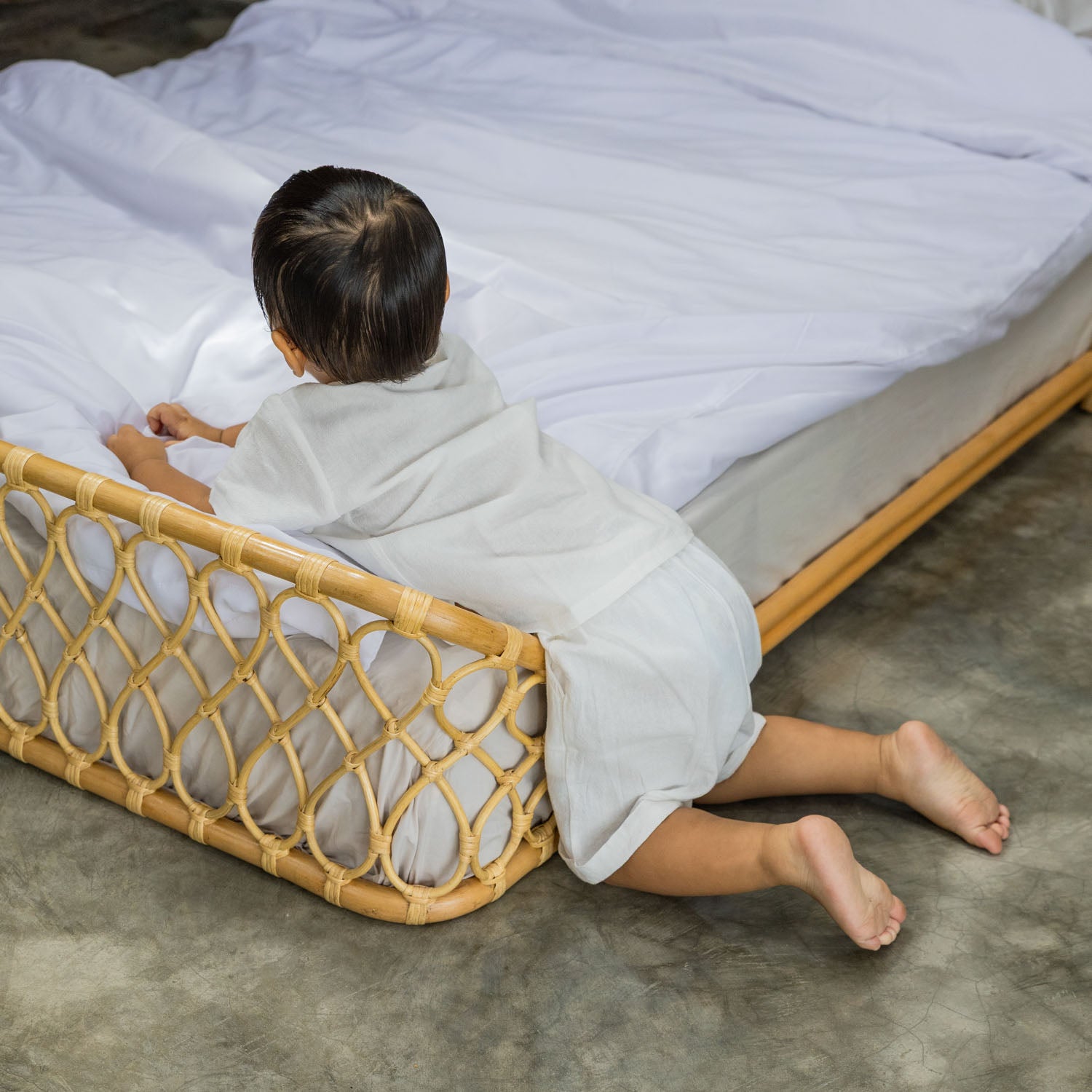Child sitting next to the handcrafted rattan wicker Layla Montessori floor bed by MOMIJI, styled in a calm indoor environment.
