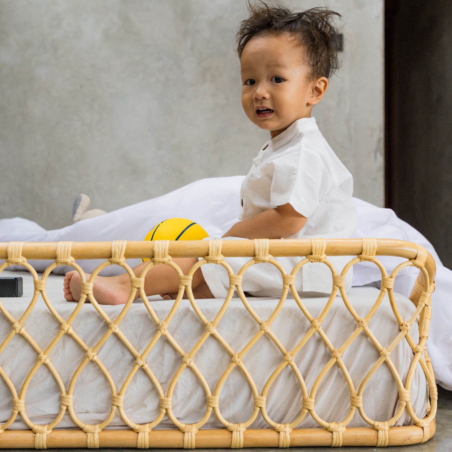Child sitting on the handcrafted Layla Montessori rattan floor bed by MOMIJI with a yellow ball, highlighting the woven rattan detailing on the frame.