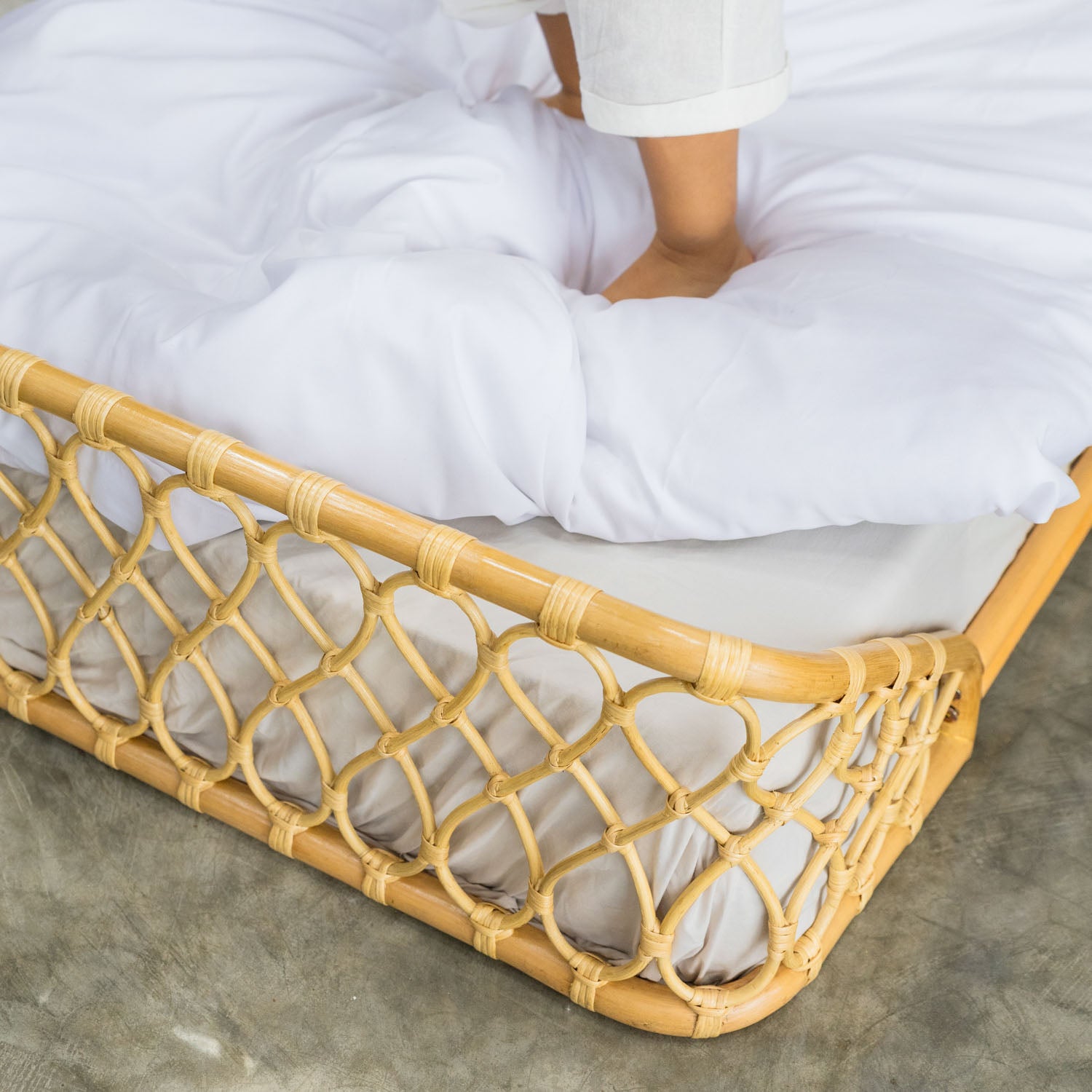 Handcrafted Layla Montessori rattan floor bed by MOMIJI in natural beige with white bedding, set on a concrete floor in a minimal indoor setting.