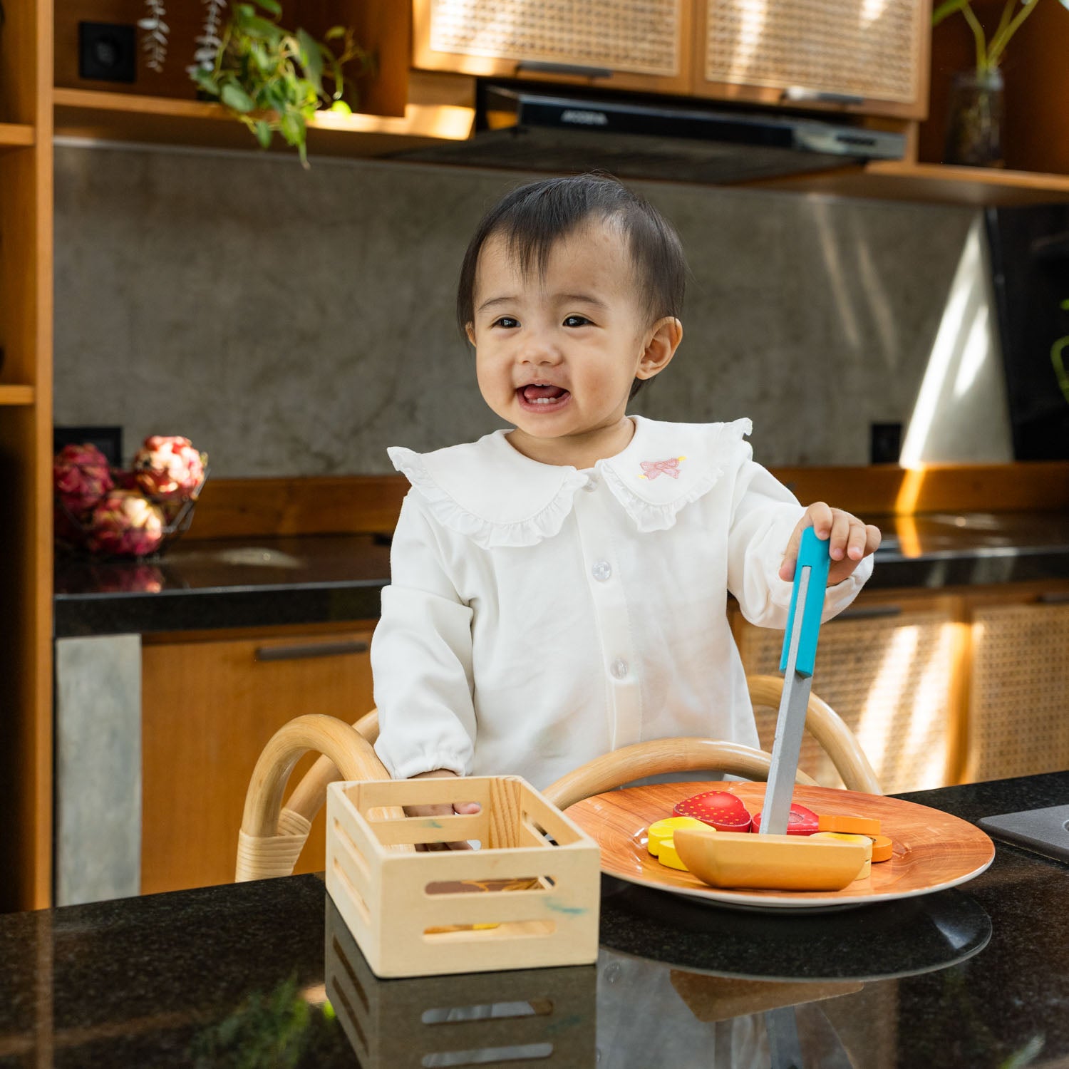 Child standing on a handcrafted Leon Montessori learning tower by MOMIJI with a rattan structure, holding a toy knife and plate in a kitchen.