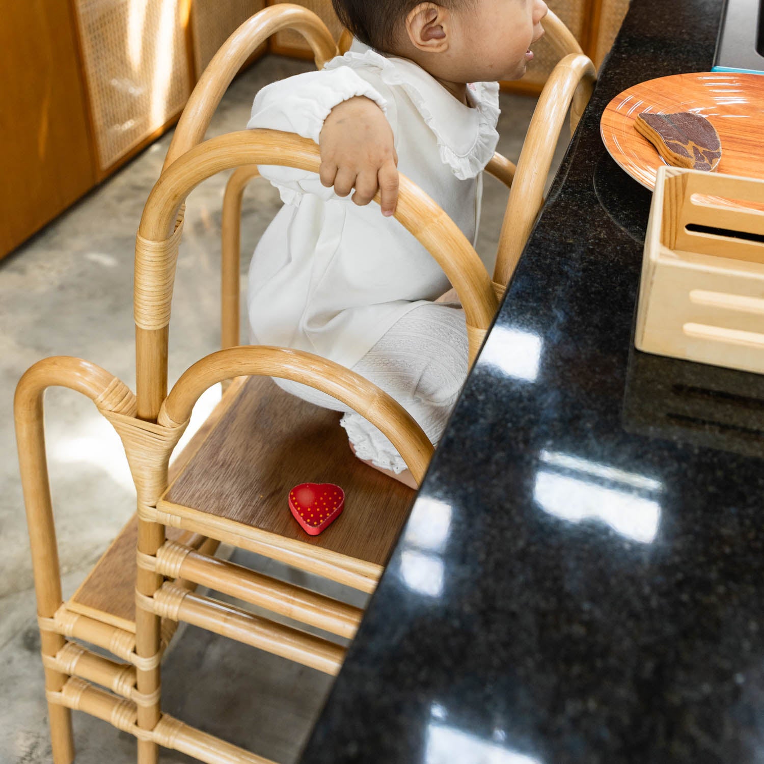 Child sitting on the handcrafted rattan wicker leon montessori learning tower by MOMIJI, holding the side support for balance.
