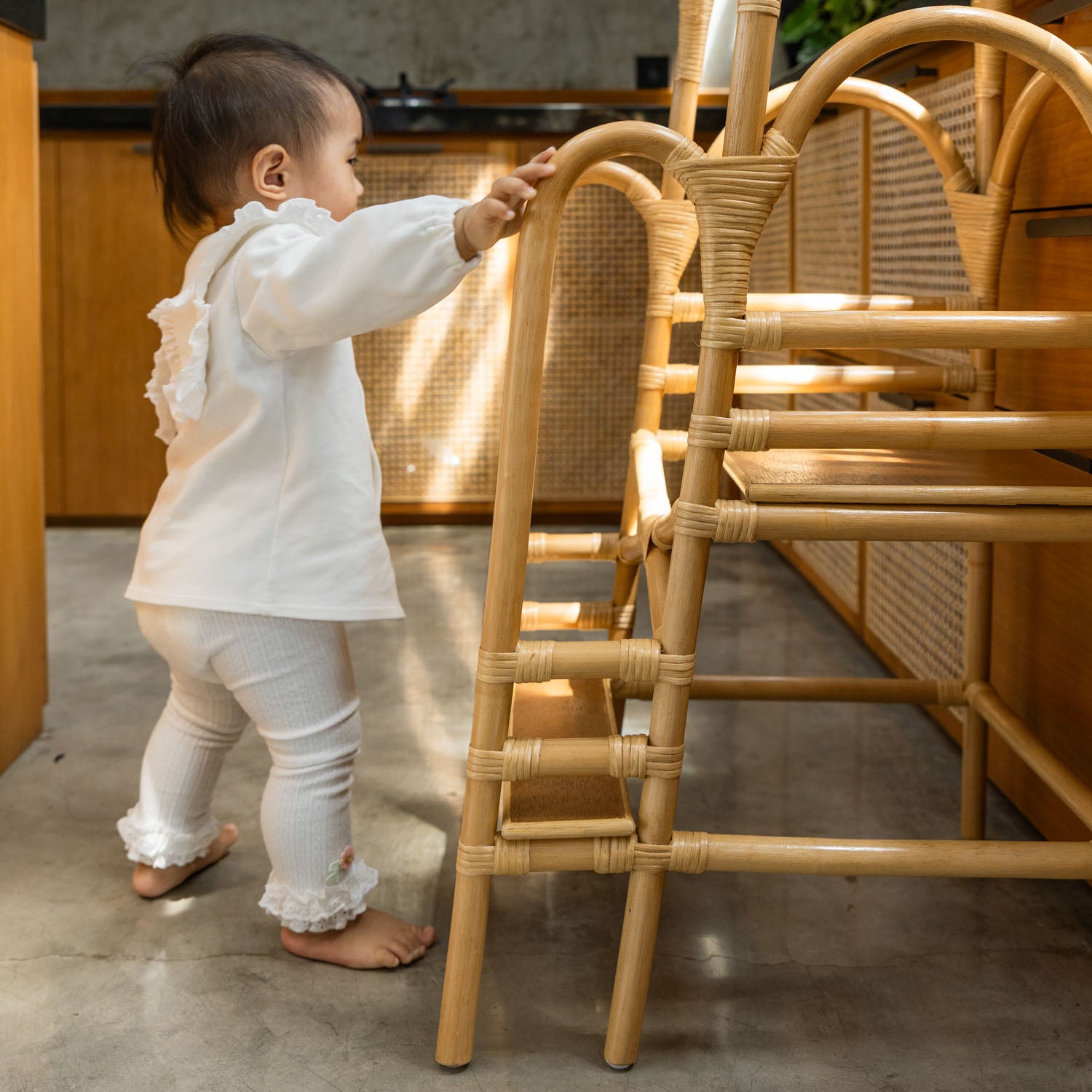 Child in a white outfit standing next to a handcrafted Leon Montessori learning tower by MOMIJI made with rattan detailing.