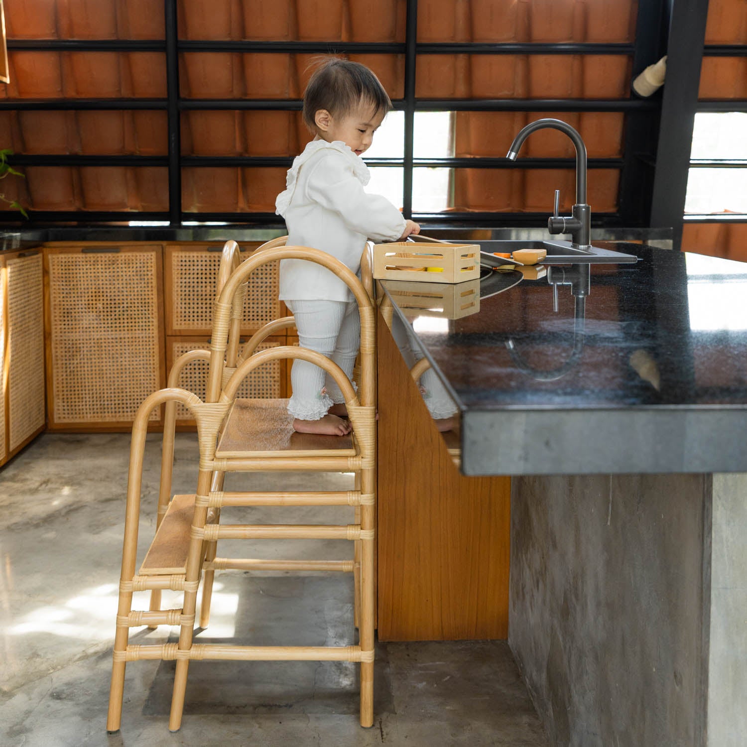 Child standing on a handcrafted Leon Montessori learning tower by MOMIJI in a modern kitchen, featuring a rattan wicker structure and wooden board with side safety supports.