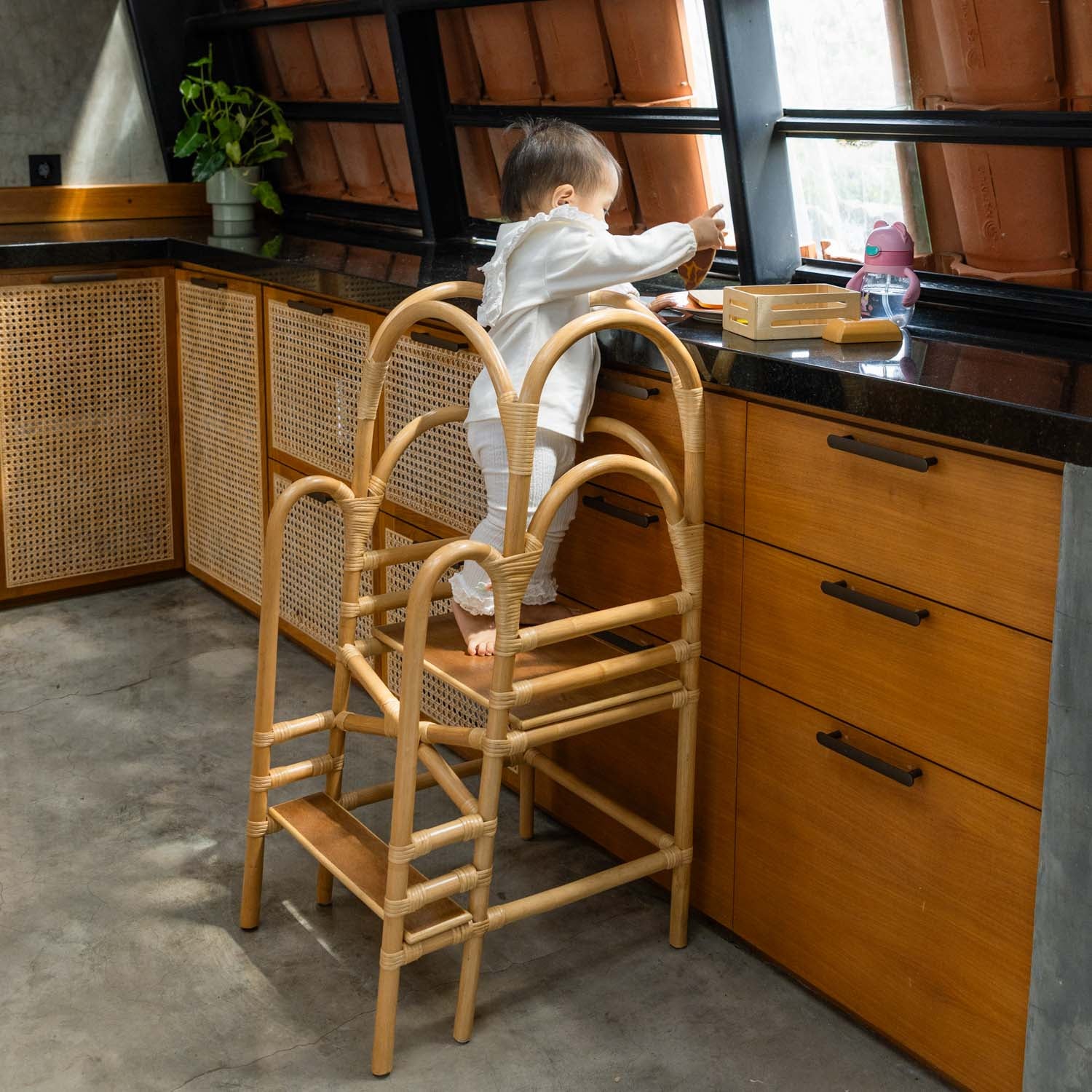 Child using a handcrafted rattan wicker leon montessori learning tower by MOMIJI to reach a kitchen counter, designed with safety supports on each side.