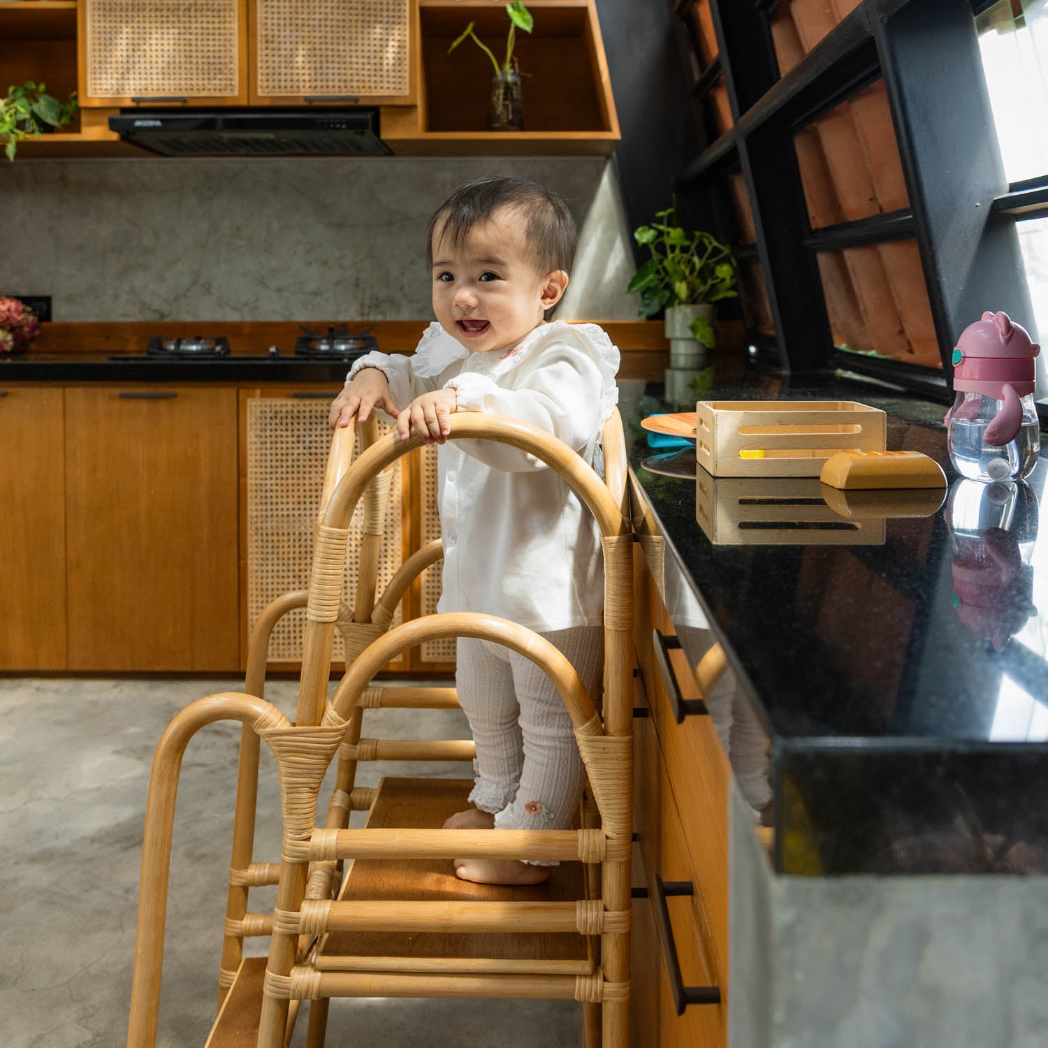 Side view of a child standing on a handcrafted Leon Montessori learning tower by MOMIJI, showing the rattan wicker frame with side safety rails.