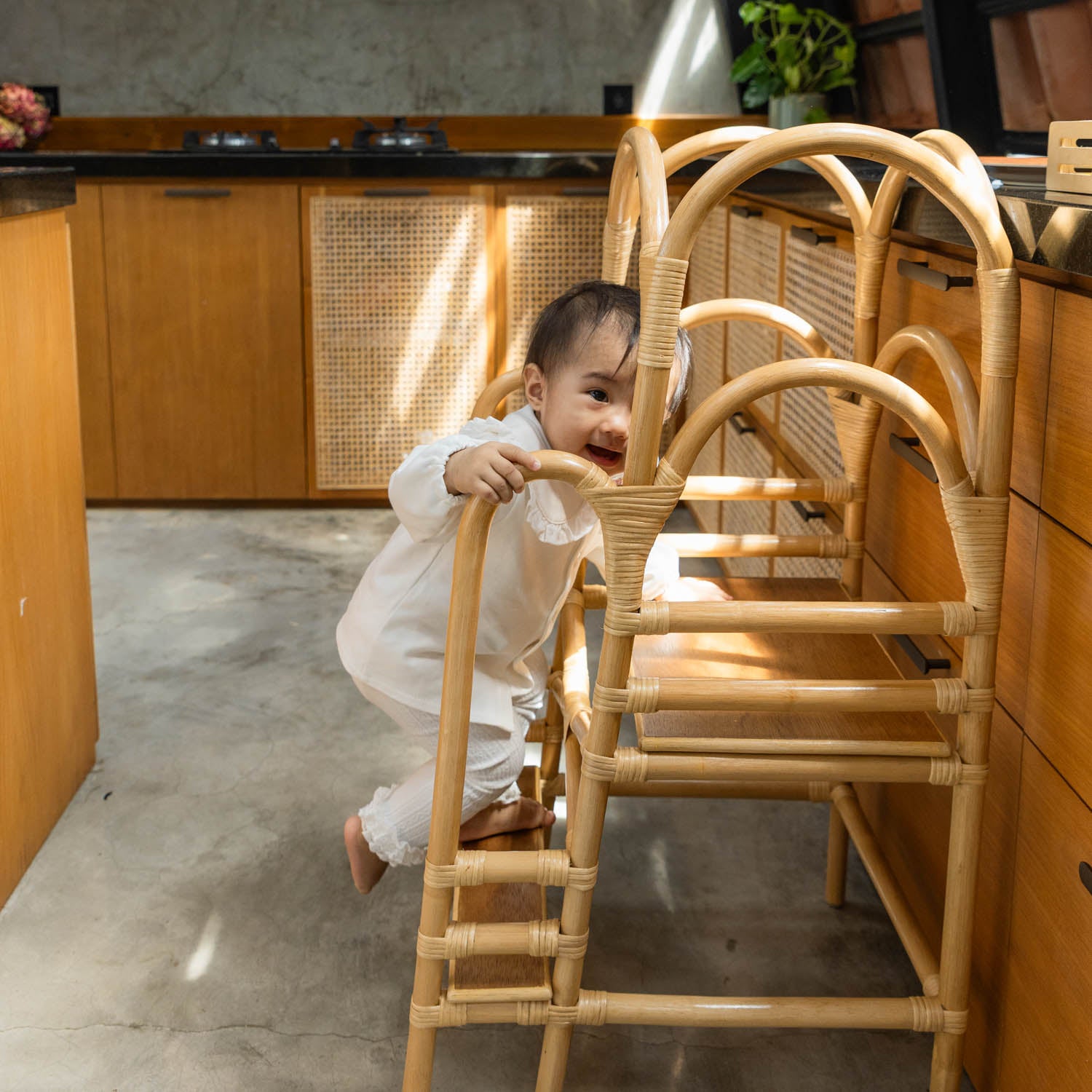 Child stepping down to the lower step of a handcrafted wicker rattan leon montessori learning tower by MOMIJI while holding the side support.