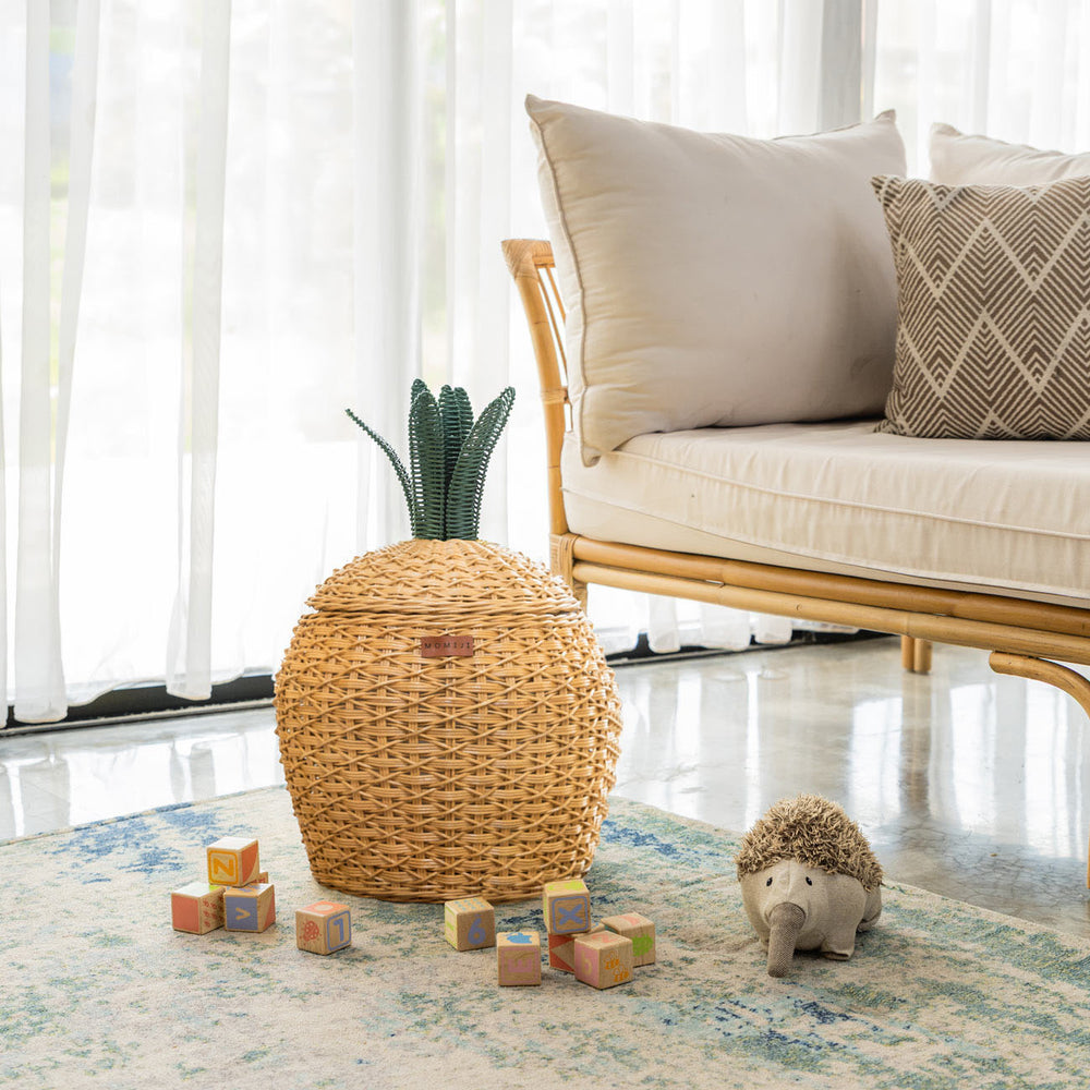 Handcrafted natural beige rattan wicker pineapple storage basket in medium size by MOMIJI, featuring a green crown lid, styled in a living room with children’s toys and a rattan bench.