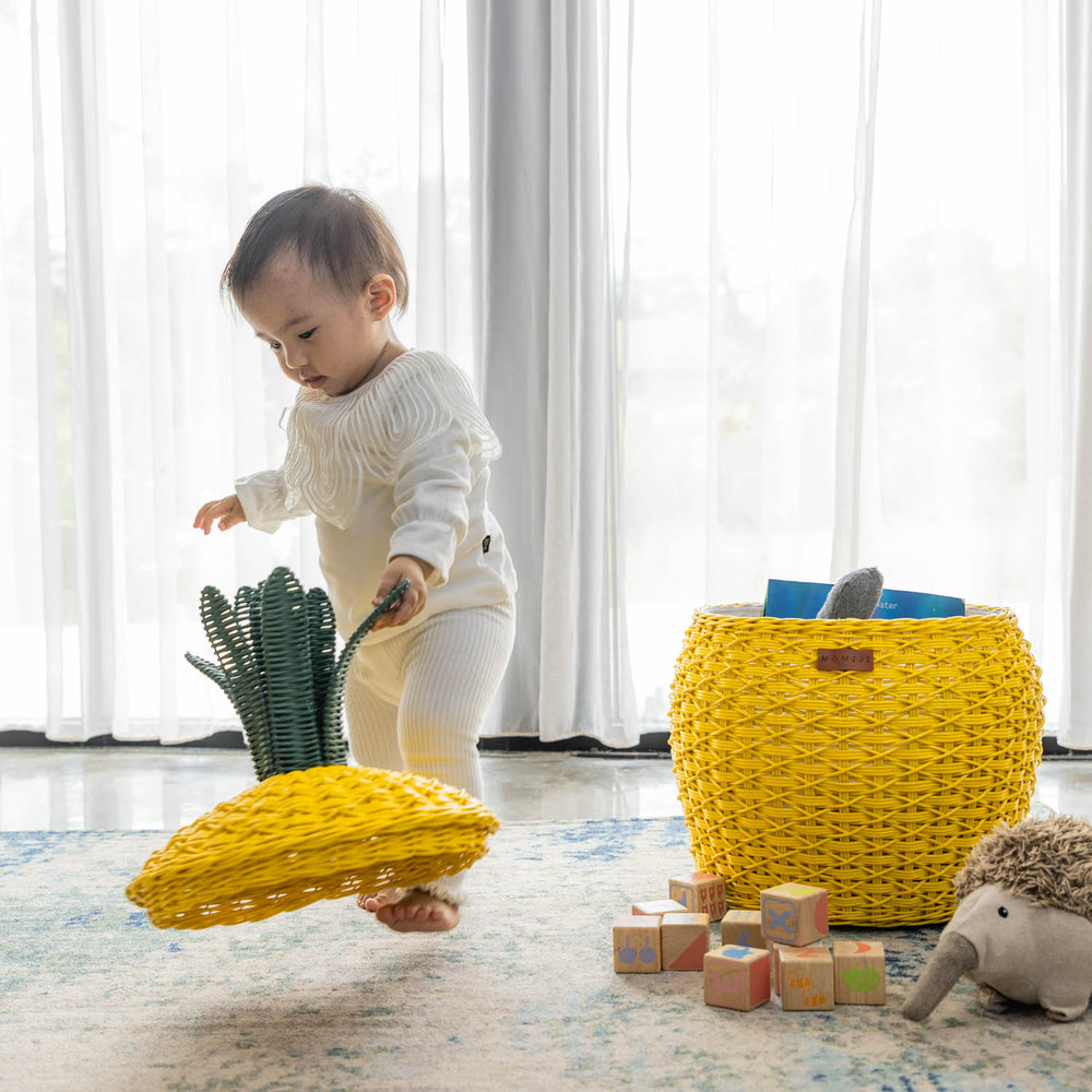 Child removing the lid from a handcrafted yellow rattan wicker pineapple storage basket by MOMIJI, styled on a rug with toys in a room with soft curtains, supporting independent play.