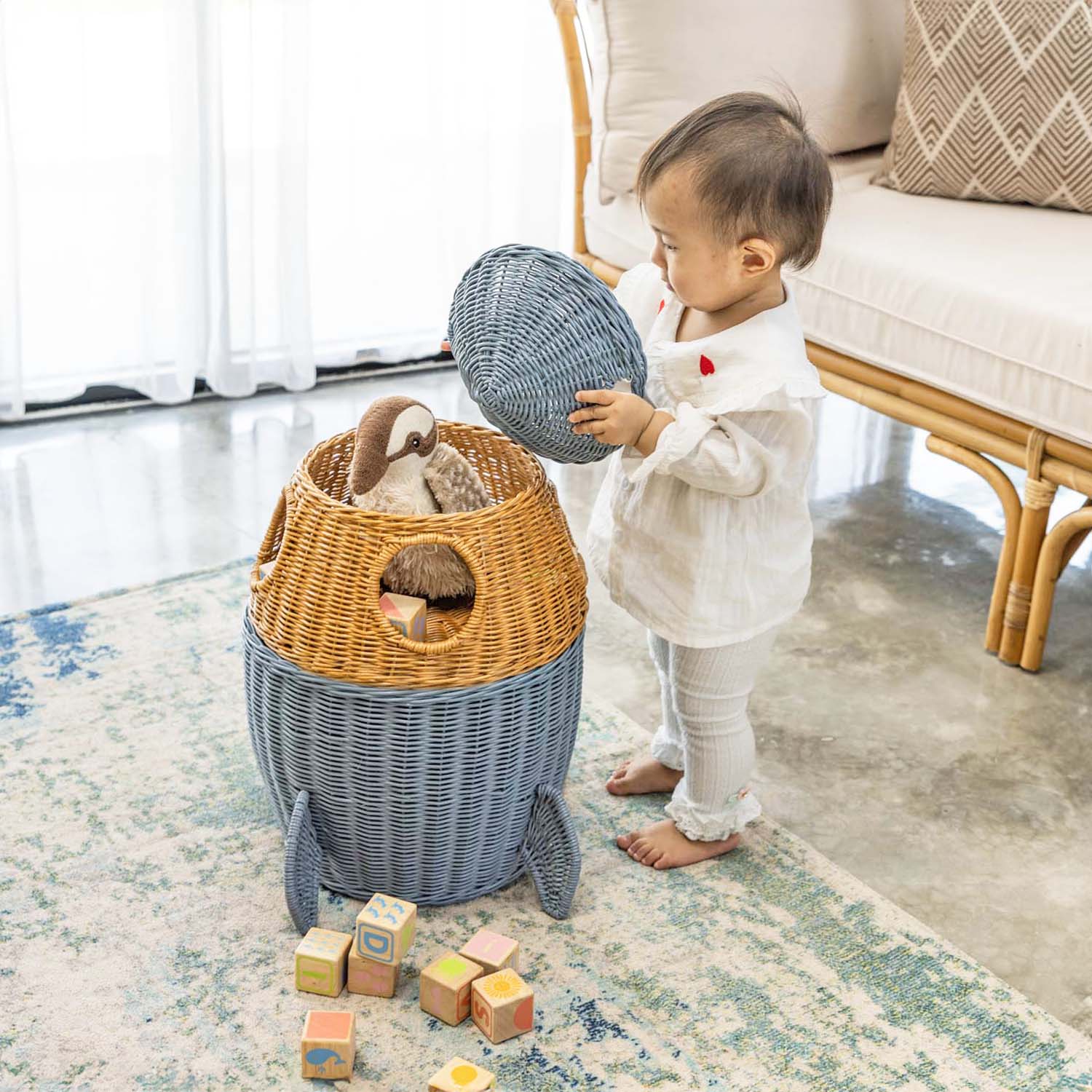 Child standing beside a handcrafted rattan wicker spaceship storage basket by MOMIJI in grey and brown, opening the lid to reveal stuffed toys, featuring rounded windows and rocket legs, styled on a rug in a living room.
