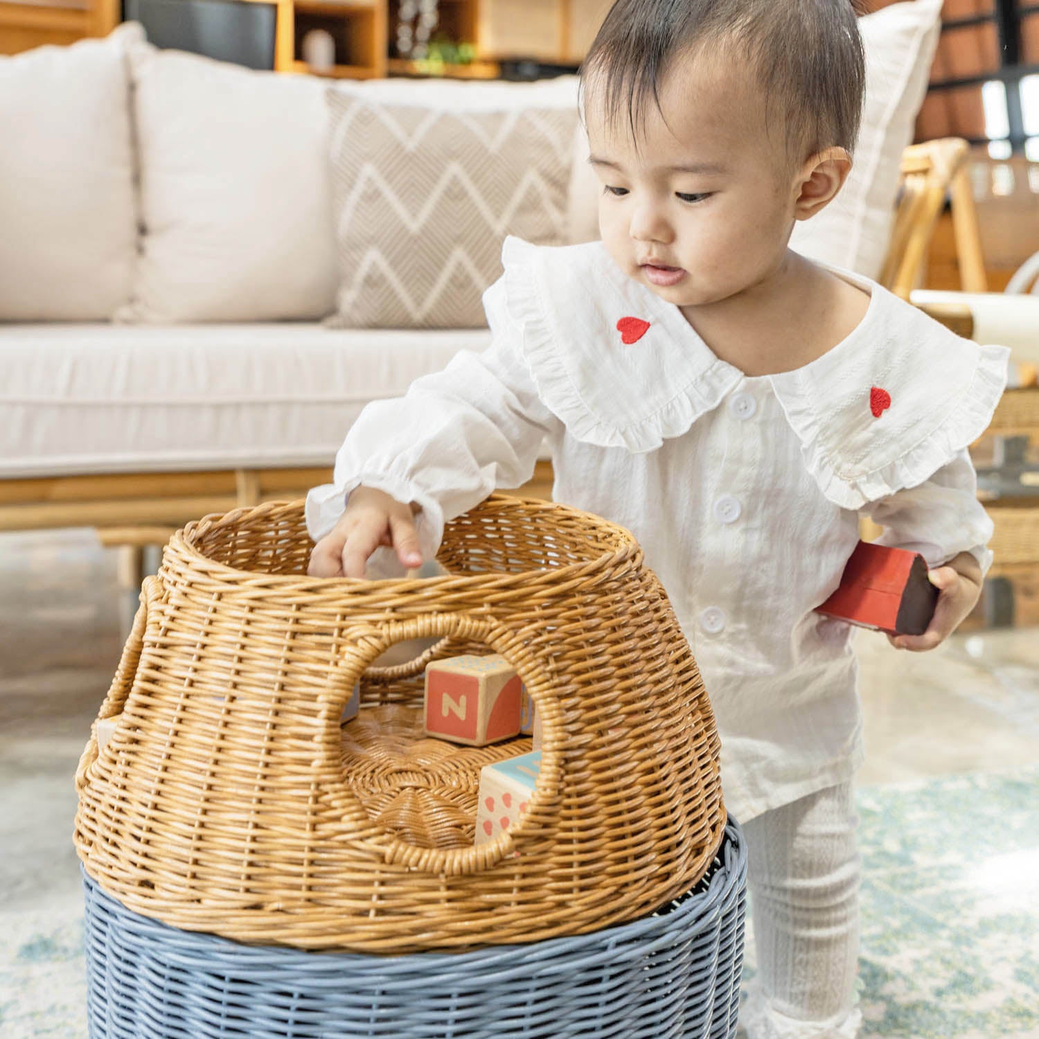 Child putting a toy inside a handcrafted rattan wicker spaceship storage basket by MOMIJI in a living room setting, featuring rounded windows.