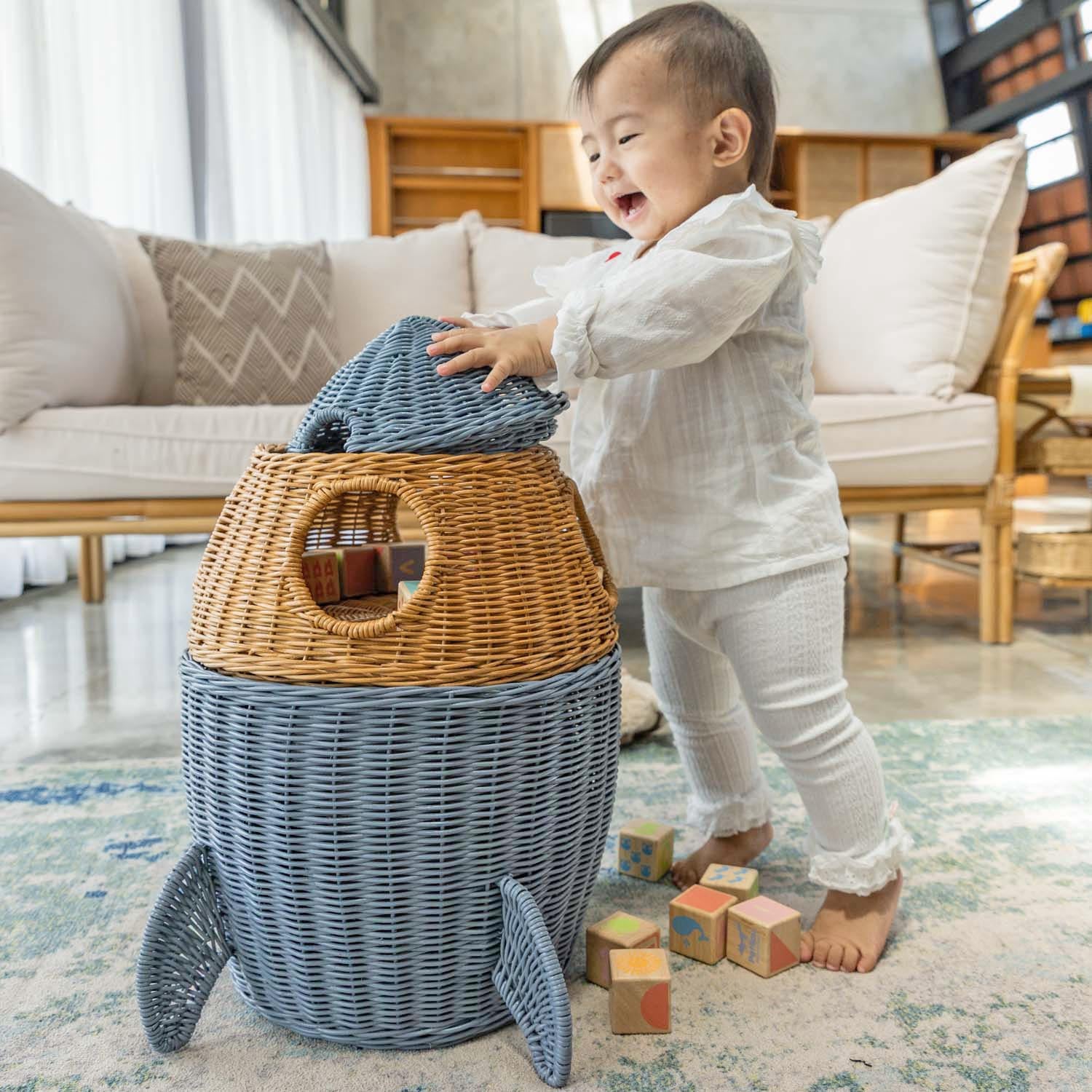 Child smiling while holding a lid of the handcrafted rattan wicker spaceship storage basket by MOMIJI in grey and brown in a living room setting, featuring rounded windows and rocket legs.