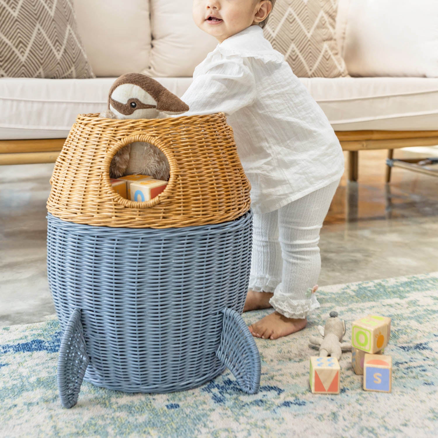 Child playing with a handcrafted rattan wicker spaceship storage basket by MOMIJI, taking toys from the opened basket in a living room, featuring rounded windows and rocket legs.