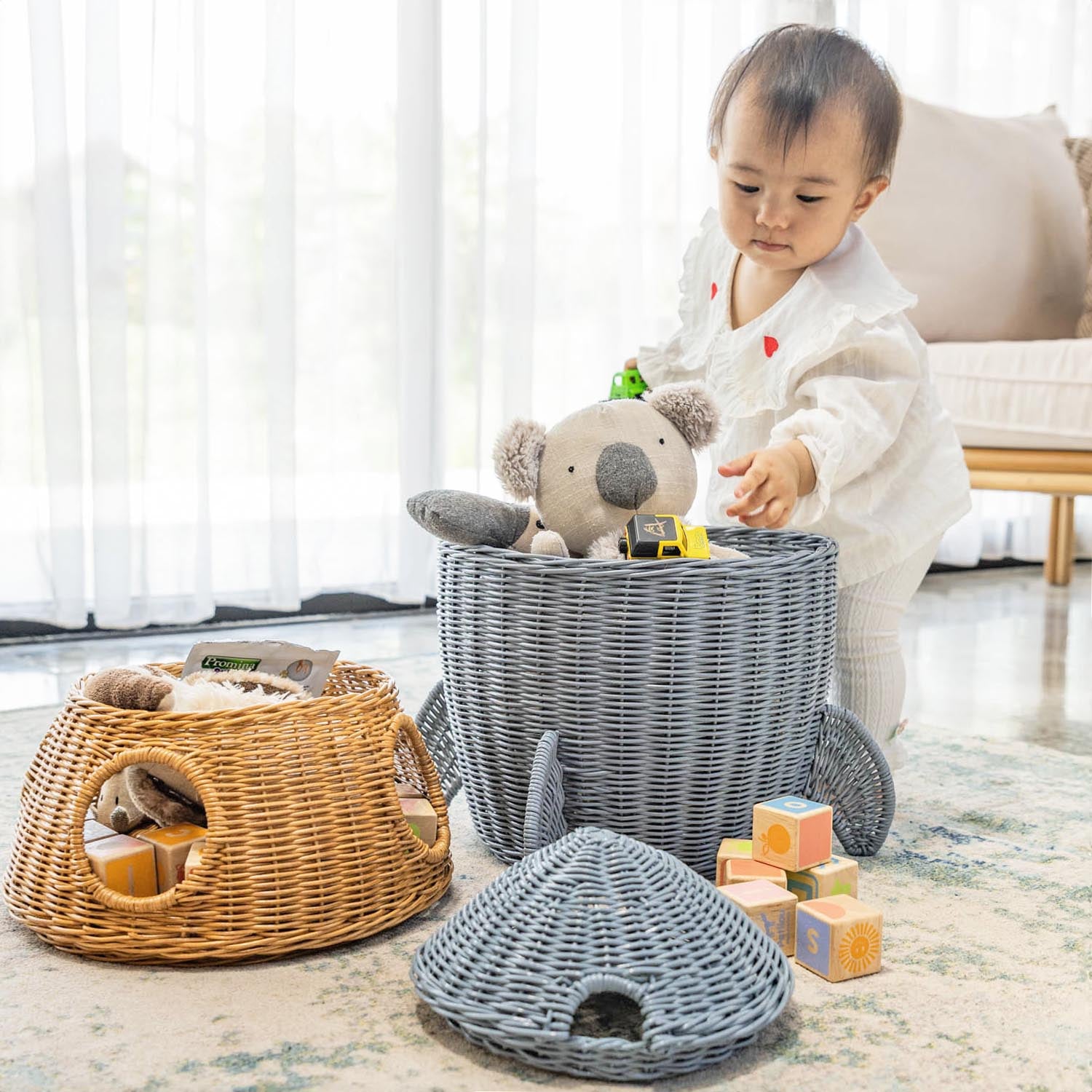 Child playing with toys from the bottom tier of a handcrafted rattan wicker spaceship storage and basket by MOMIJI, middle tier and the lid placed beside it on a rug near a large window.