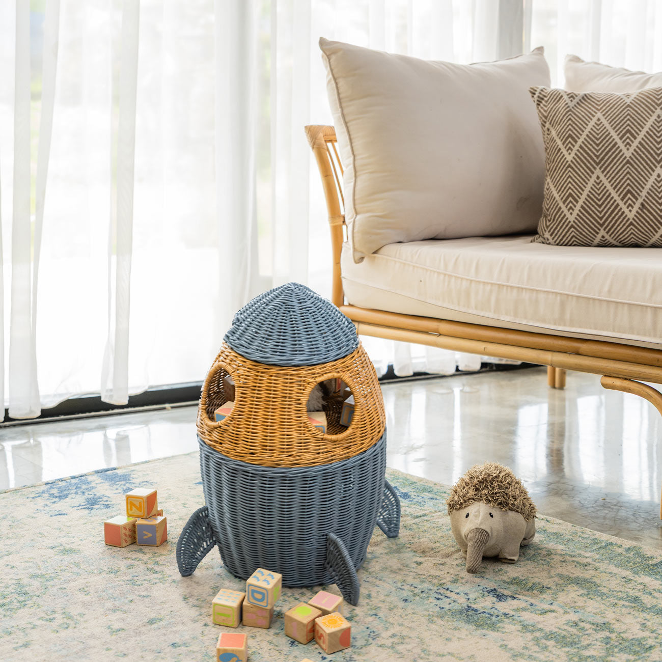 Handcrafted rattan wicker spaceship 3 tier storage basket by MOMIJI in grey and brown, featuring round window details and rocket legs, filled with children’s toys on a rug with colorful blocks and a plush hedgehog in a kids room with a rattan bench.