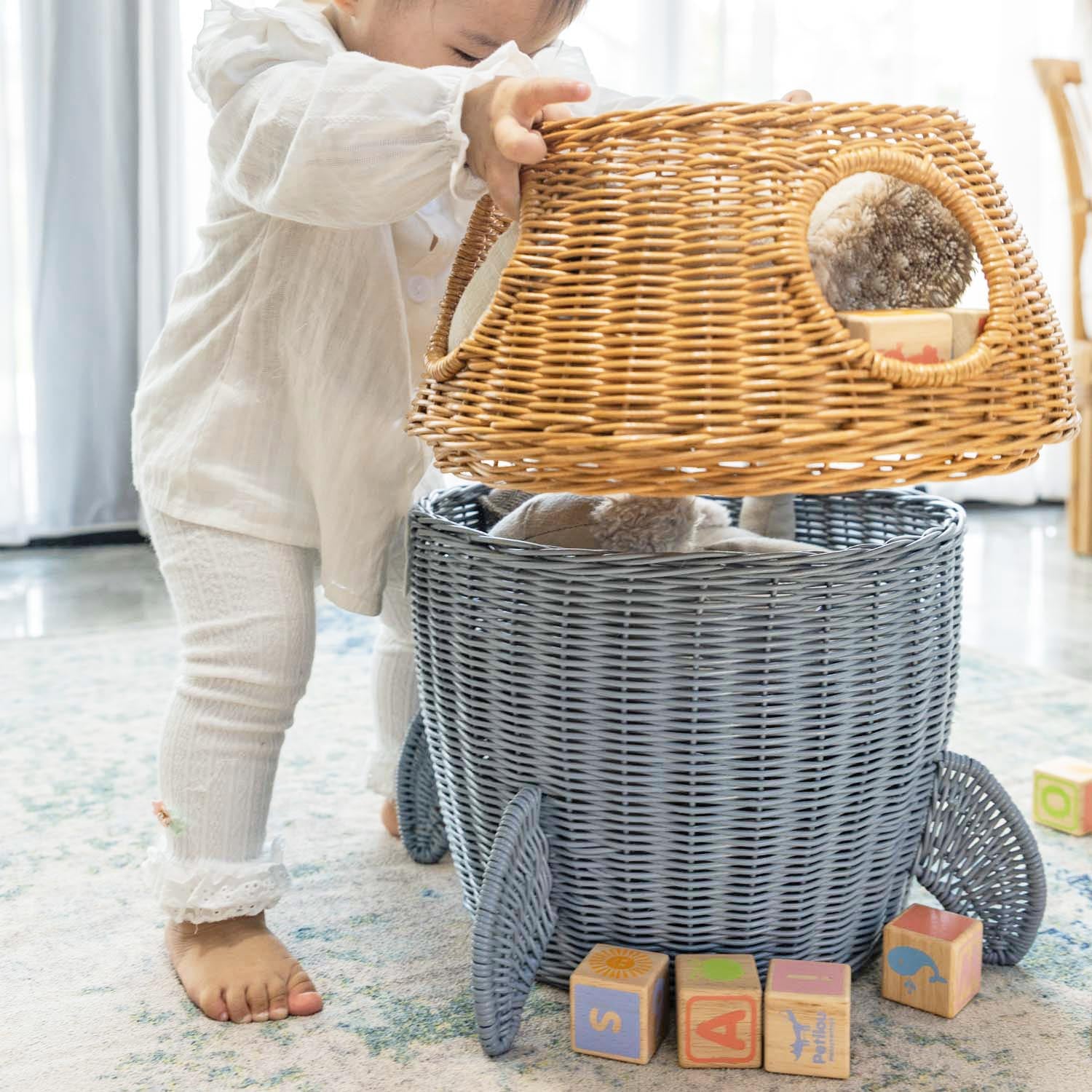 Child removing the middle tier of a handcrafted rattan wicker spaceship storage basket by MOMIJI and opening the bottom tier filled with toys, featuring rounded windows and rocket legs.