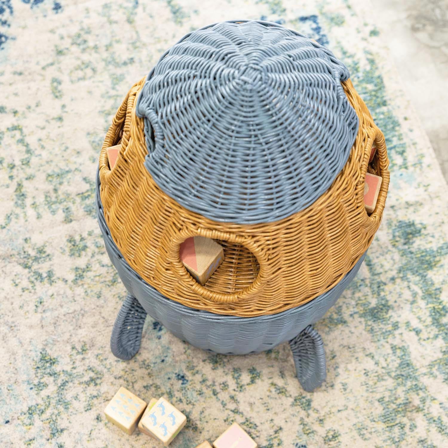 Top view of a handcrafted rattan wicker spaceship storage basket by MOMIJI on a carpet with colorful blocks nearby, featuring rounded windows and rocket legs.