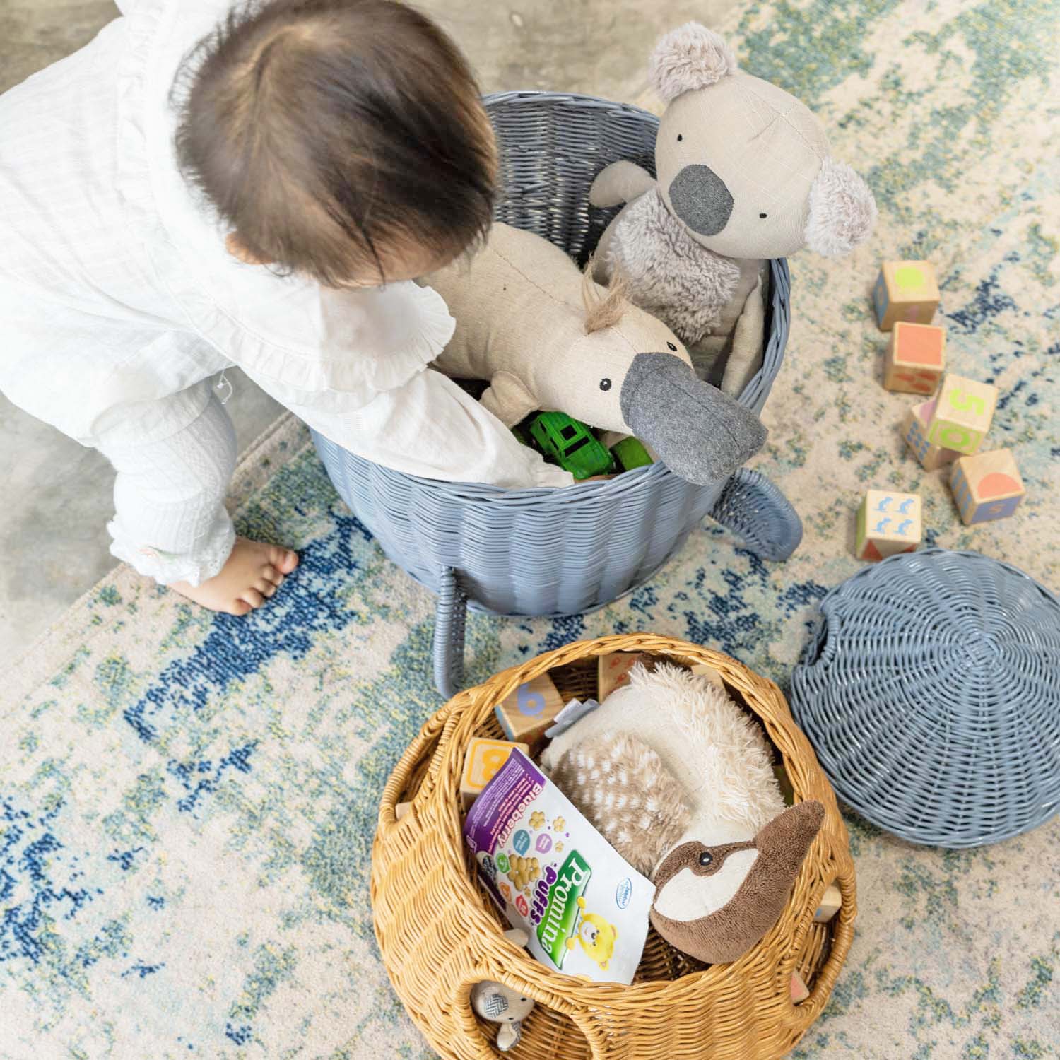 Top view of a child taking toys from the bottom tier of the handcrafted rattan wicker spaceship storage and basket by MOMIJI, with the middle tier and lid placed beside it on a patterned rug.