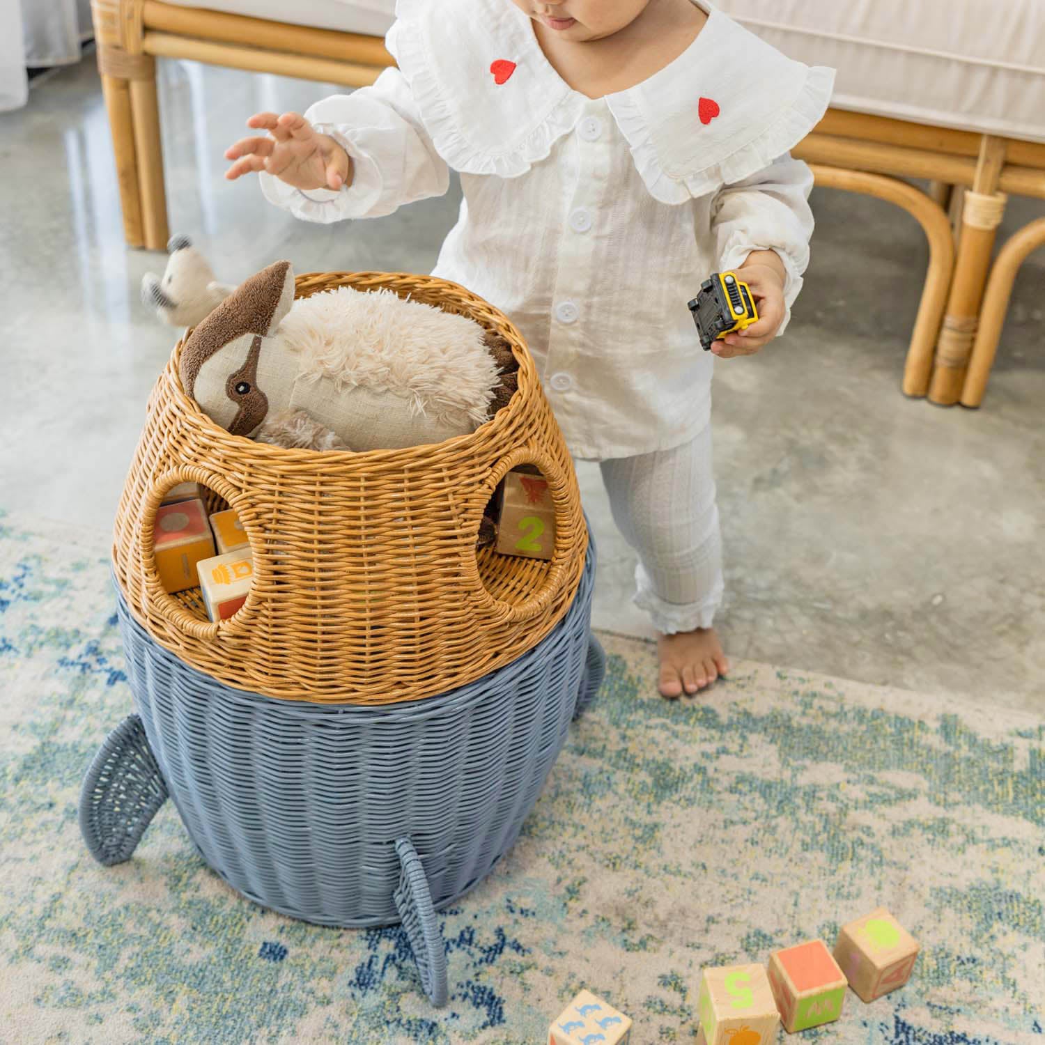 Child holding a toy car next to an opened handcrafted rattan wicker spaceship storage basket by MOMIJI filled with toys on a rug, featuring rounded windows and rocket legs.
